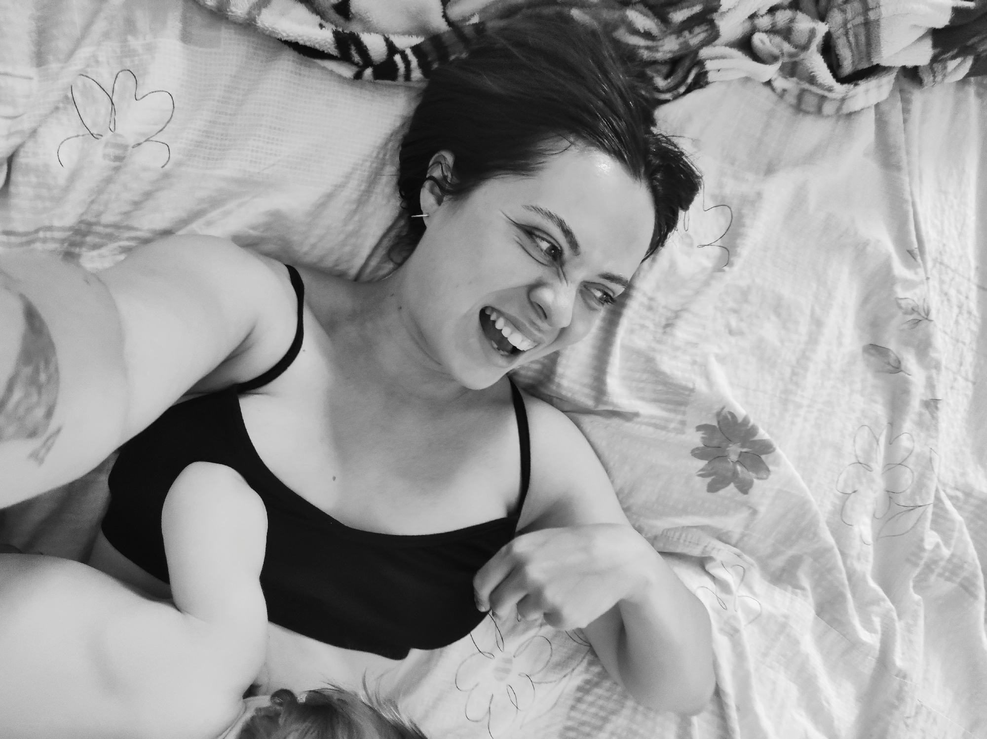 A black and white, high-angle shot depicts a woman lying on a patterned bed or blanket, with part of a baby visible in the lower-left corner. The woman is wearing a dark-colored, spaghetti-strap top and is smiling, displaying her teeth. Her hair is dark and appears damp. Part of her arm is visible with a tattoo. The bed or blanket has a floral design and a striped blanket near the top of the frame. The location and specific city are unknown.