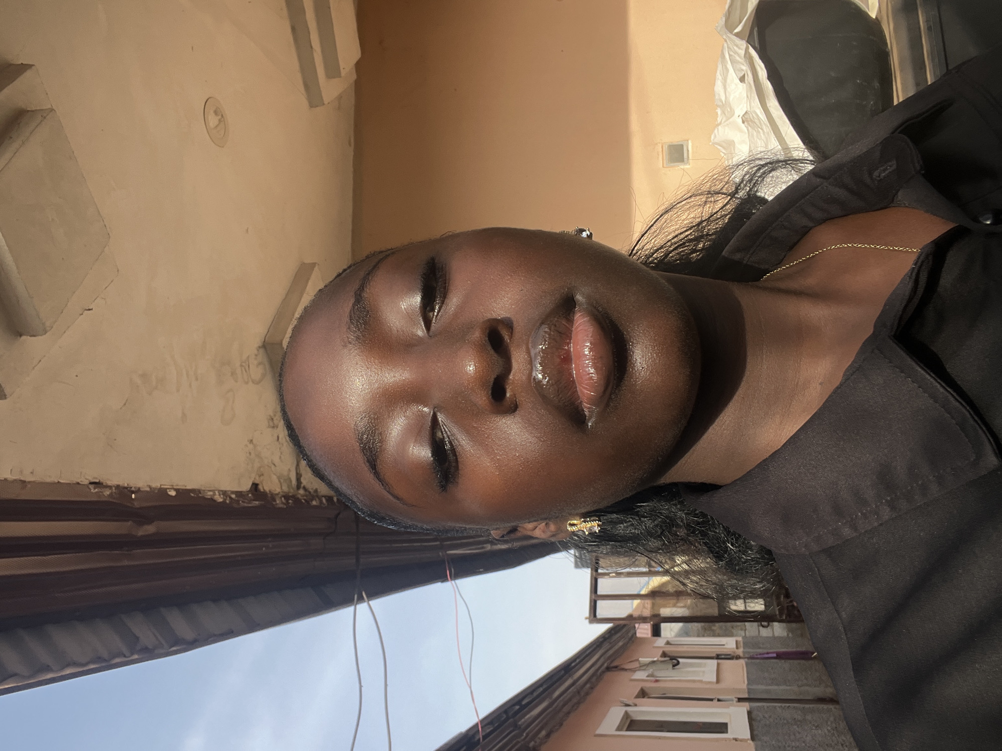 This close-up portrait features a person with dark skin, oriented vertically in the provided image but depicting the subject in a horizontal, relaxed pose. The individual's eyes are closed, and their lips are slightly parted and glossy, reflecting strong, direct sunlight. Their dark hair is visible around their face and shoulders. They are wearing a black collared shirt or top, adorned with a thin gold chain around their neck and a small, intricate golden earring in their right ear.
The scene is brightly lit by natural light, indicating it is daytime. The sun creates prominent highlights on the person's forehead, nose, and lips, emphasizing their skin's texture. The background consists of a light-colored wall, possibly beige or tan, featuring architectural details such as raised sections or moldings. Below this, a portion of a corrugated roof structure is visible, against a backdrop of a clear blue sky. Some thin wires are also present in the lower part of the frame. The setting appears to be outdoors or in an open, semi-enclosed area, likely within an urban or suburban environment. There is no visible text in the image.