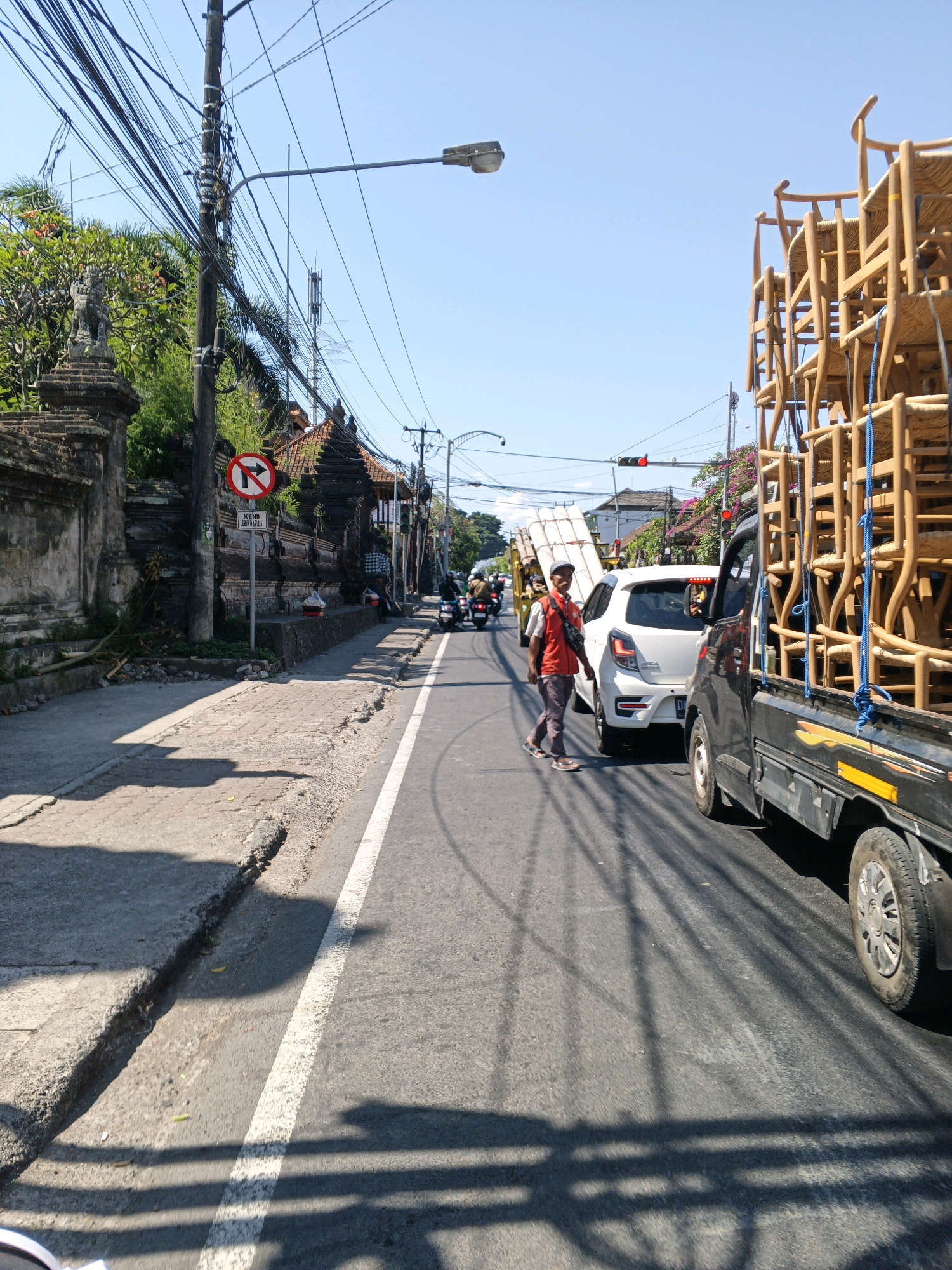 The image depicts a street scene in Abiansemal, Indonesia, on a clear, sunny day. A black flatbed truck heavily laden with stacked wooden chairs occupies the right side of the frame, its cargo secured with blue rope. A white car is positioned to the left of the truck, also on the road. A man in an orange vest and a red shirt is walking across the road in front of the white car, appearing to be directing traffic or overseeing the movement of vehicles. In the background, a variety of buildings with traditional Indonesian architecture are visible, along with a prominent traffic light displaying a red signal. Numerous utility poles with a dense network of wires run along the street. A "No Right Turn" sign with Indonesian text below is posted on a pole on the left side of the road, near a stone wall. The road has a white dividing line. The scene is bustling with activity, suggesting a commercial area or a busy thoroughfare within the town. The presence of motorcycles and the general street environment reinforce the location as a populated urban or semi-urban area.