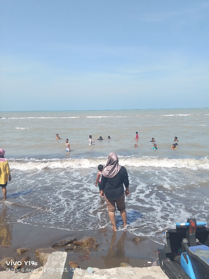 This image captures a daytime coastal scene in Limbangan, Indonesia. A clear, light blue sky occupies the top third of the frame, meeting a flat horizon. The ocean water is a light brown color, characterized by moderate waves breaking towards the shore. Approximately 10 to 12 individuals, mostly children and young adults, are present in the water, wading or swimming; some are partially submerged, with only their heads visible above the surface.
In the foreground, a woman wearing a light-colored hijab, a black long-sleeved shirt, and brown pants stands in the shallow surf, facing away from the camera and towards the ocean. A child in a white and red striped top stands directly in front of her, also facing the water. To the far left, another individual in a yellow top and dark shorts is partially visible, standing in the water near the shoreline. The immediate shoreline consists of dark, wet sand, interspersed with several rocks and concrete fragments. A dark wooden boat or structure with blue accents is partially visible in the bottom right corner of the frame. A digital watermark in the lower left corner states "VIVO Y19s Akbhar & Arvan. 12 Okt 2025, 13:44."