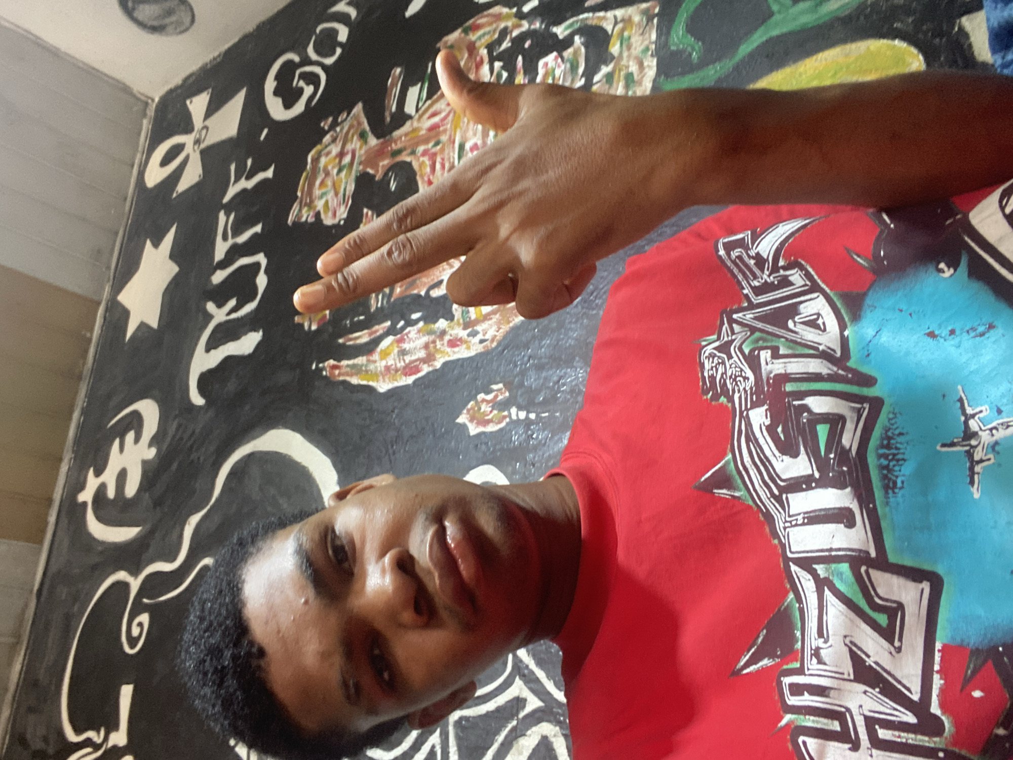 A person is posing for a photo. The person is wearing a red t-shirt with a graphic design. The person is holding their hand up in a gesture, potentially mimicking a gun shape. The background is a wall with artwork, appearing to be a mural. The artwork includes stylized script and other decorative elements. The location appears to be in Lekki, Nigeria.