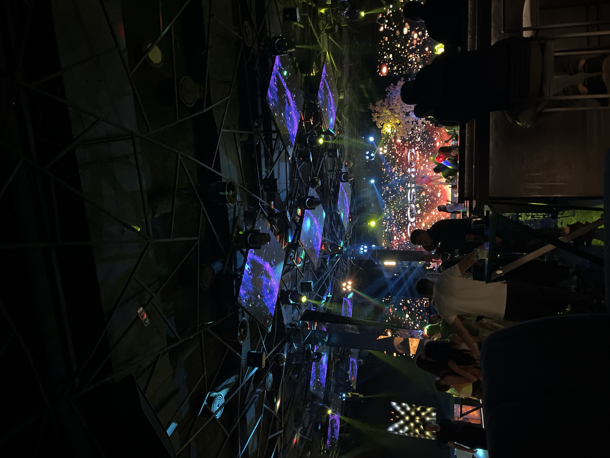The image captures a night scene at a club or event in Plovdiv, Bulgaria. In the background, there is a large stage or installation composed of triangular or diamond-shaped screens, illuminated with vibrant colors and displaying abstract or space-themed imagery. Spotlights and stage lighting are also present, adding to the visual spectacle.
Foregrounded are numerous people, some standing at tables or seated, suggesting they are patrons of the establishment. Many are obscured or partially visible due to the angle of the shot. The overall atmosphere is lively, indicative of a bustling nightlife venue. The architecture suggests a modern or industrial design, with metal beams and geometric patterns forming part of the structural elements of the space.
