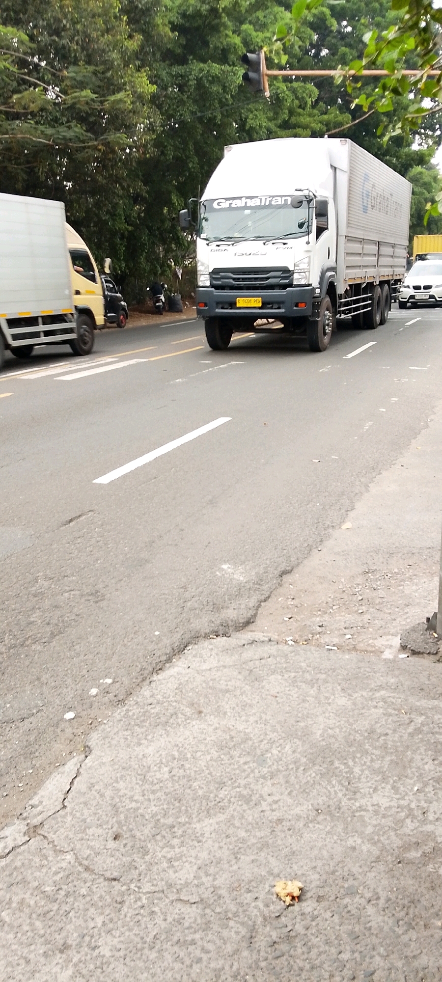The image shows a street scene in Depok, Indonesia, likely on a cloudy day given the lighting. In the foreground, a portion of a cracked asphalt road is visible, with some debris and cigarette butts on the side. A white truck with the branding "Graha Trans" is prominent in the midground, driving on the road. This truck is a delivery truck. To the left of the white truck, a yellow and white box truck is partially visible, also on the road. Further back, a black motorcycle is parked on the side of the road, next to some black trash bags. In the background, lush green trees line the street, with a traffic light visible above the white truck. A white car is also seen in the distance to the right. The overall environment suggests a typical urban or suburban street in Indonesia, with traffic and common street elements.