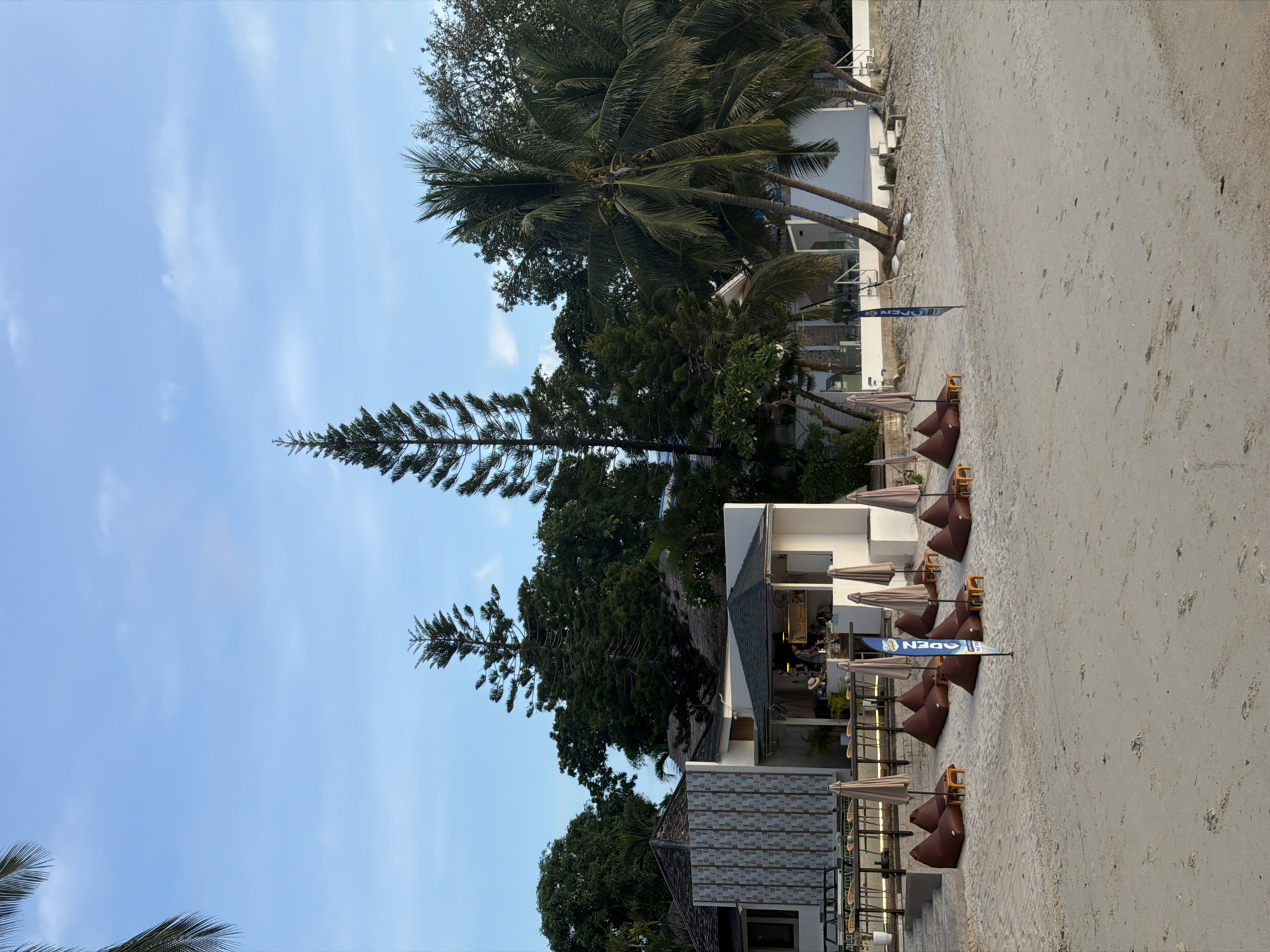This media file depicts a tropical beach scene in Baan Bang Rak, Thailand, likely during the late afternoon or early evening given the soft lighting. The image is oriented vertically, showing a stretch of sandy beach leading towards a row of buildings and lush greenery.
The main subjects include several beach umbrellas and beanbag chairs arranged on the sand, suggesting a seating area for a beachside establishment. Palm trees and other tropical foliage are prominent, framing the scene and adding to the exotic atmosphere. In the background, modern white buildings with glass elements and outdoor seating can be seen, indicating a resort or restaurant.
A blue flag with white text is planted in the sand, displaying the word "OPEN". There are no visible people in the frame, implying the establishment may be about to open or has just closed for the day, or that the photo was taken during a quiet period. The sky is a clear blue with a few wispy clouds. The sand appears undisturbed except for some footprints. The overall mood conveyed is one of relaxation and tropical tranquility.
