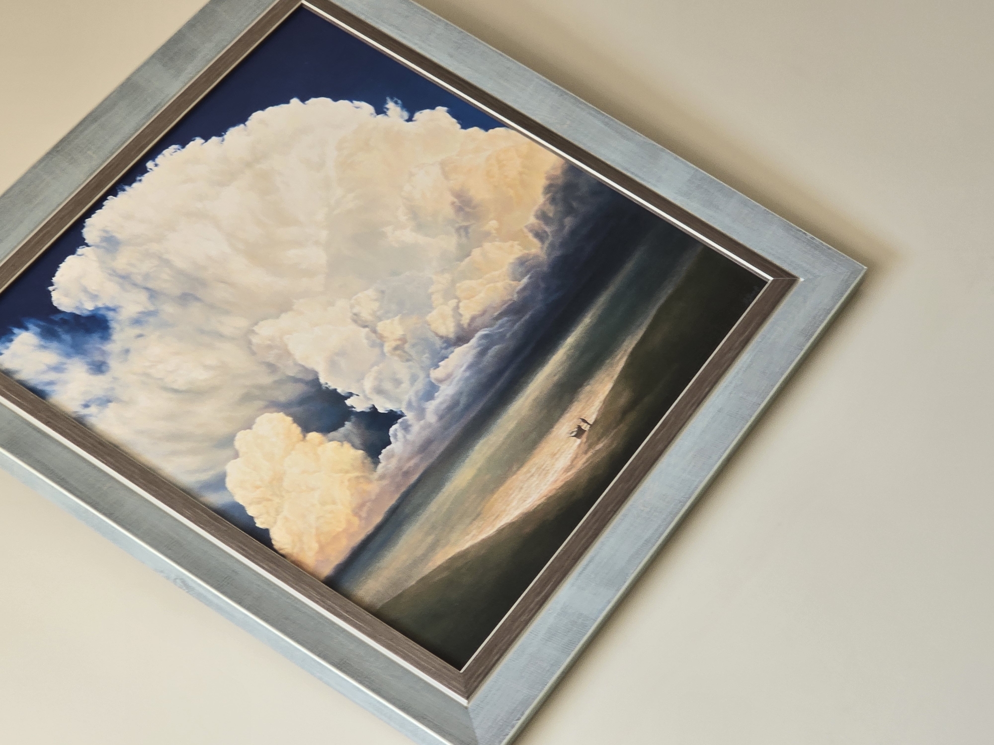 A framed painting hangs on a light beige wall. The painting depicts a large, billowing cloud formation against a blue sky. The clouds transition into a land or sea surface, with two small figures standing in the distance. The frame is a double frame, the inner part being a wooden color and the outer part being a lighter, metallic, silver/blue. The painting is oriented at an angle to the viewer.