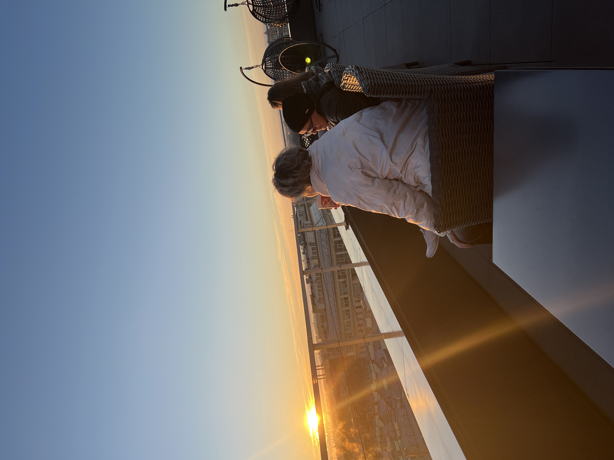 Two people are seated in wicker chairs on a high balcony, overlooking a city at sunset. The setting sun casts a warm glow over Saint Petersburg, Russia, visible in the distance. One person wears a white jacket, while the other is dressed in dark clothing and a hat. They appear to be engaged in conversation, enjoying the view. The balcony features a glass barrier and dark tiles, with additional outdoor seating visible in the background.