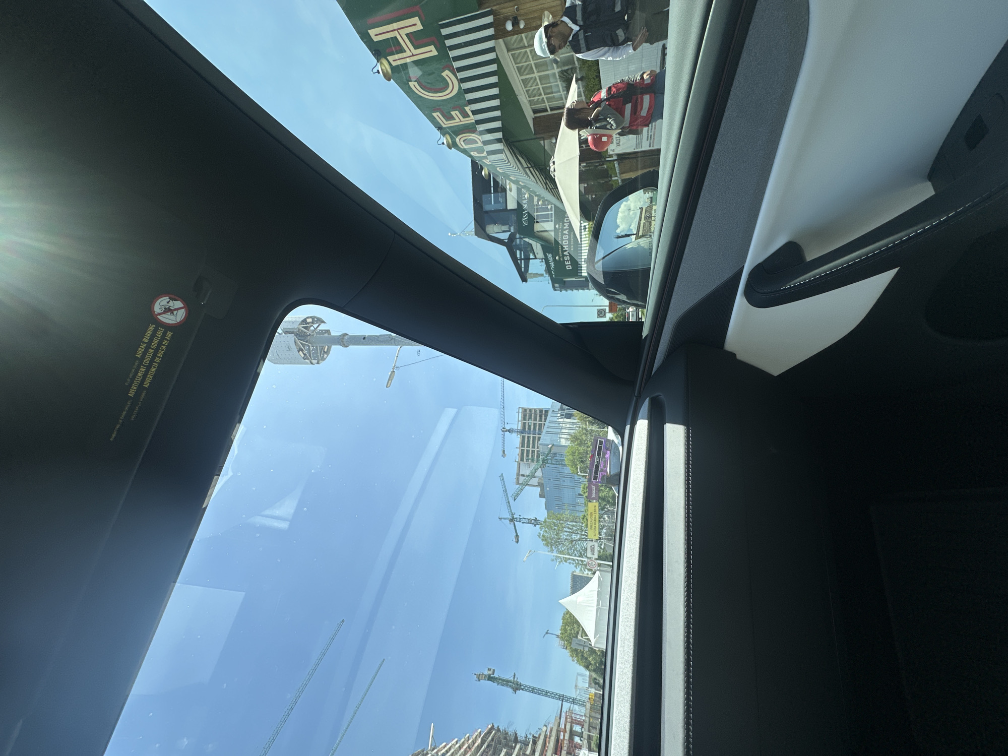 The image is taken from the interior of a vehicle, looking out through the windshield and side window. Outside, a bright, sunny day is visible with a clear blue sky. In the distance, several construction cranes tower over unfinished buildings, indicating an active construction site within a city. The architecture suggests an urban environment.
To the right, a building with a green facade and the word "CRECH" partially visible is present. Two individuals are standing in front of this building. One person is wearing a red vest and a red hard hat. The other person is wearing a white hard hat and a dark vest over a white shirt. They appear to be interacting with someone or observing the surroundings.
A white umbrella-shaped structure, possibly a tent, is situated in the middle ground. The interior of the car shows a black dashboard and a white and black door panel with a silver handle. A sun visor is visible at the top of the windshield, with a warning sticker indicating "AIRBAG WARNING" and "AVERTISSEMENT DE BOLSA DE AIRE."
Based on the presence of the "CRECH" signage and the architectural style, this scene is likely located in the Angelópolis area of Puebla, Mexico. The construction cranes suggest ongoing urban development.