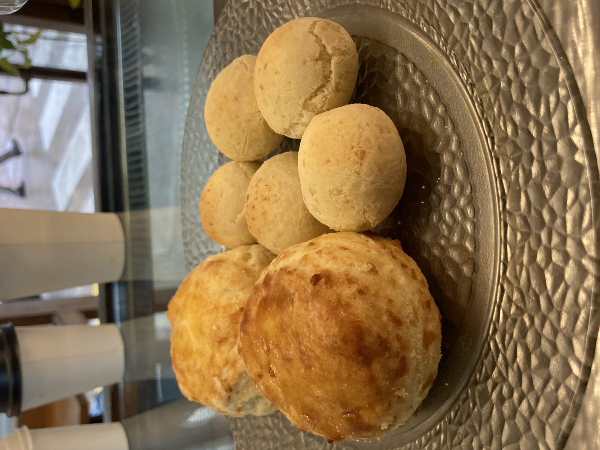 The image displays a close-up, top-down shot of several golden-brown baked goods, likely cheese breads (pão de queijo), arranged on a decorative, textured plate. There are approximately nine individual pieces, varying slightly in size and color. The smaller, lighter-colored pieces are stacked towards the top and center of the plate, while two larger, more deeply golden-brown pieces with a slightly crispier appearance are positioned at the bottom left. The surfaces of the baked goods show some cracks and irregularities typical of fresh baking.
The plate itself is round, appearing to be made of a metallic or metallic-finished material, and features a prominent, textured pattern resembling small, interconnected facets or hammered dimples across its surface and rim.
The background is intentionally blurred, creating a shallow depth of field that keeps the food in sharp focus. Despite the blur, elements of an indoor setting are discernible. These include vertical lines that could be parts of window frames or display shelves, and some reflective surfaces, possibly glass, in front or around the food. In the left background, out-of-focus white cylindrical objects, possibly cups or dispensers, are visible, further suggesting a cafe, bakery, or food service environment. The lighting appears to be ambient indoor light, highlighting the textures of the food and the plate. No people or visible text are present in the image. The image is a still life focused on the presentation of the baked goods.