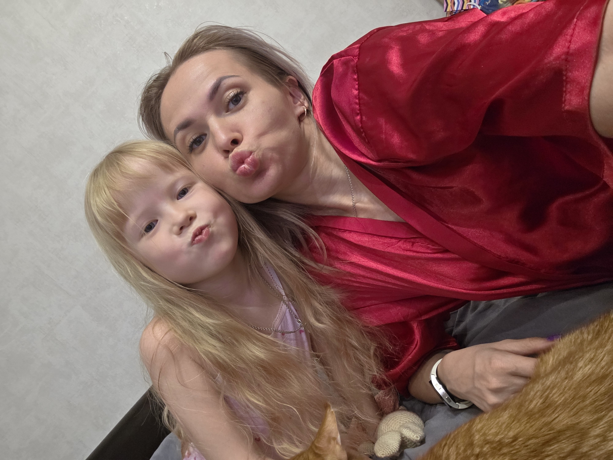 A woman and a child, both with pursed lips, are in a selfie taken indoors. The woman, who is wearing a red robe, is slightly behind and to the right of the child. The child has long blonde hair and is wearing a pink top. Part of a cat and a stuffed animal are in the bottom right corner. A grey wall is in the background. The location is unknown.