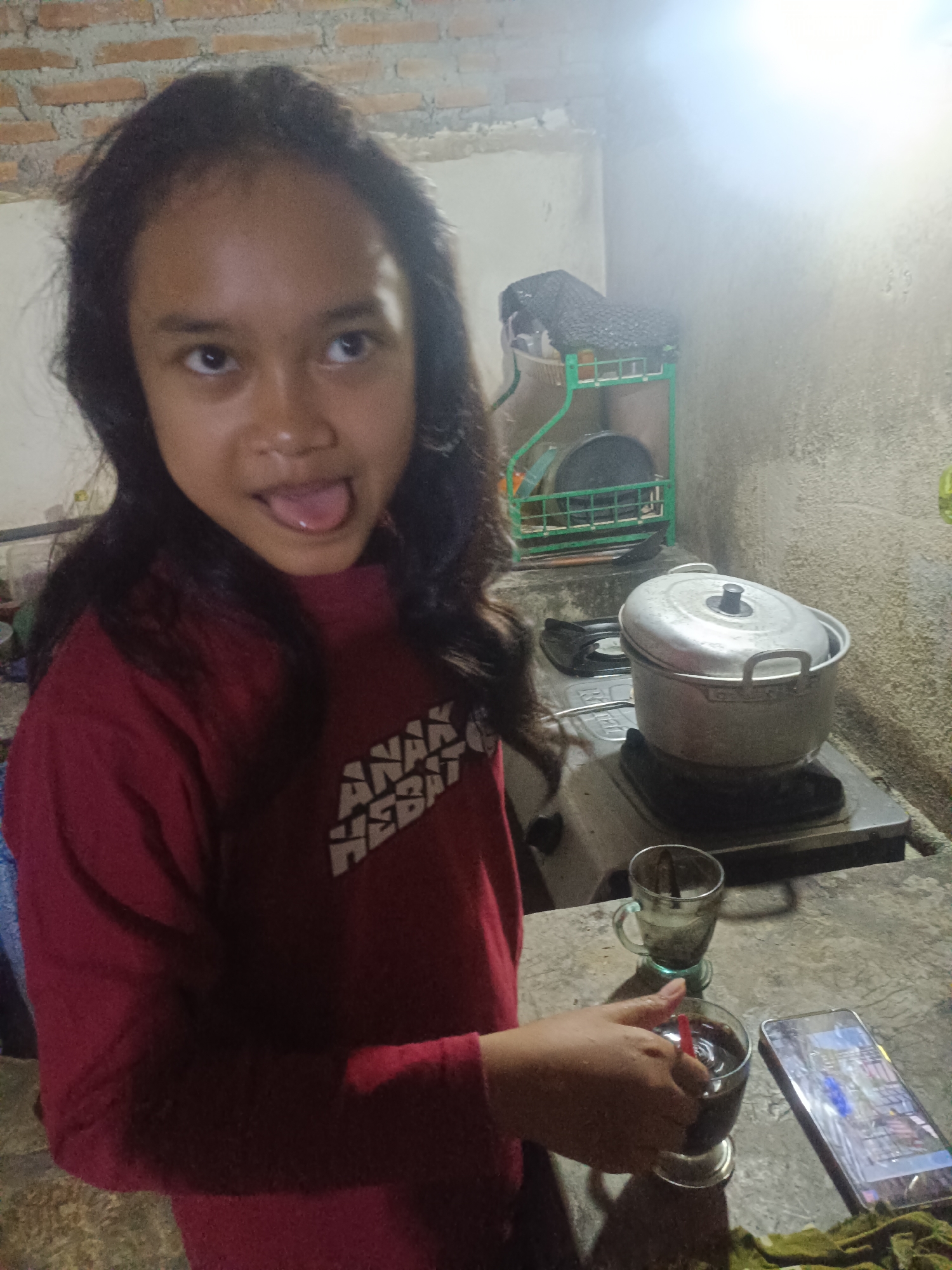 A young girl with long, dark hair sticks her tongue out playfully while looking at the camera. She is wearing a red long-sleeved shirt with white text on it that reads "ANAK HEBAT," which translates to "great child" in Indonesian. She is in a kitchen area with exposed brick walls and a concrete countertop.
On the countertop, there is a gas stove with a pot on it. Next to the stove are two glasses; one is empty and the other contains a dark liquid, likely coffee, with a red straw in it. A smartphone lies on the countertop next to the glasses. The scene appears to be indoors, possibly in a home kitchen, given the casual setting and the presence of kitchen appliances. The lighting suggests it could be evening or indoors with artificial light. The overall mood is lighthearted and playful, conveyed by the girl's expression. The location context is Tambakromo, Indonesia.