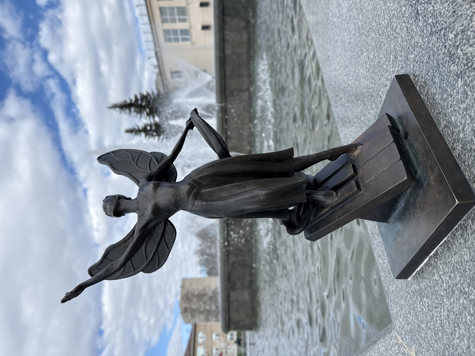 The image shows a bronze statue of a winged woman in Chaikovsky, Russia. The statue is positioned in front of a fountain with water jets visible in the background. The statue's base is mounted on a grey granite structure. The sky is cloudy with patches of blue. Buildings and trees are visible in the background, blurred by the fountain's spray.