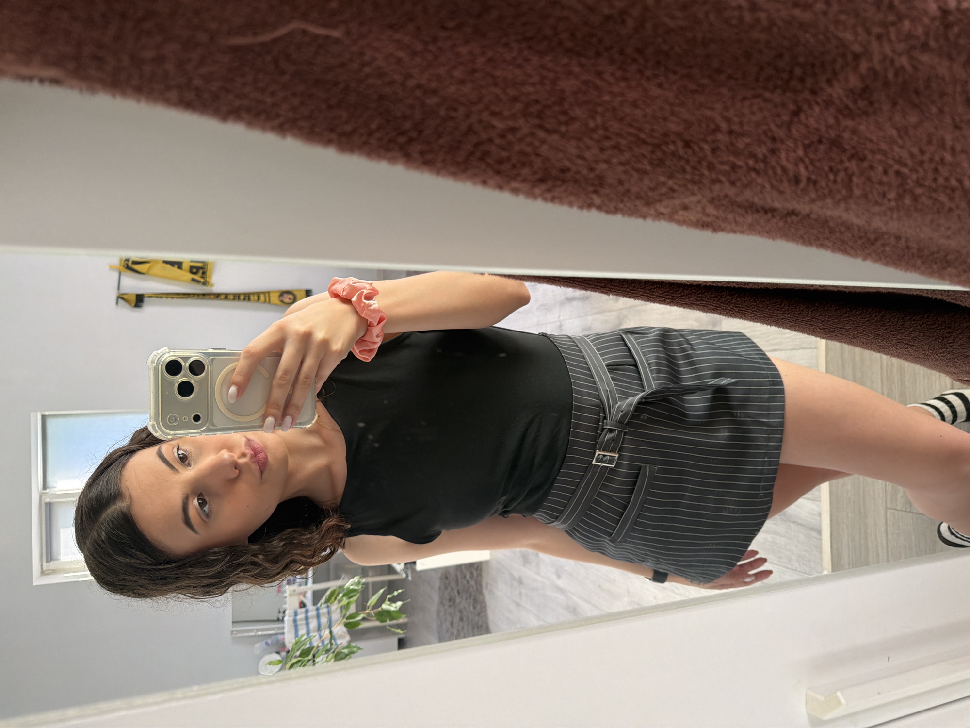A young woman with curly brown hair takes a mirror selfie in what appears to be a bedroom or dressing area. She is wearing a black sleeveless top and a dark gray pinstripe skirt with a belted waist. Her right hand is holding a smartphone, and a pink scrunchie is visible on her left wrist. In the reflection of the mirror, a window and some plants are visible in the background, along with two yellow and black scarves hanging on the wall. A brown textured towel hangs to the right of the mirror.