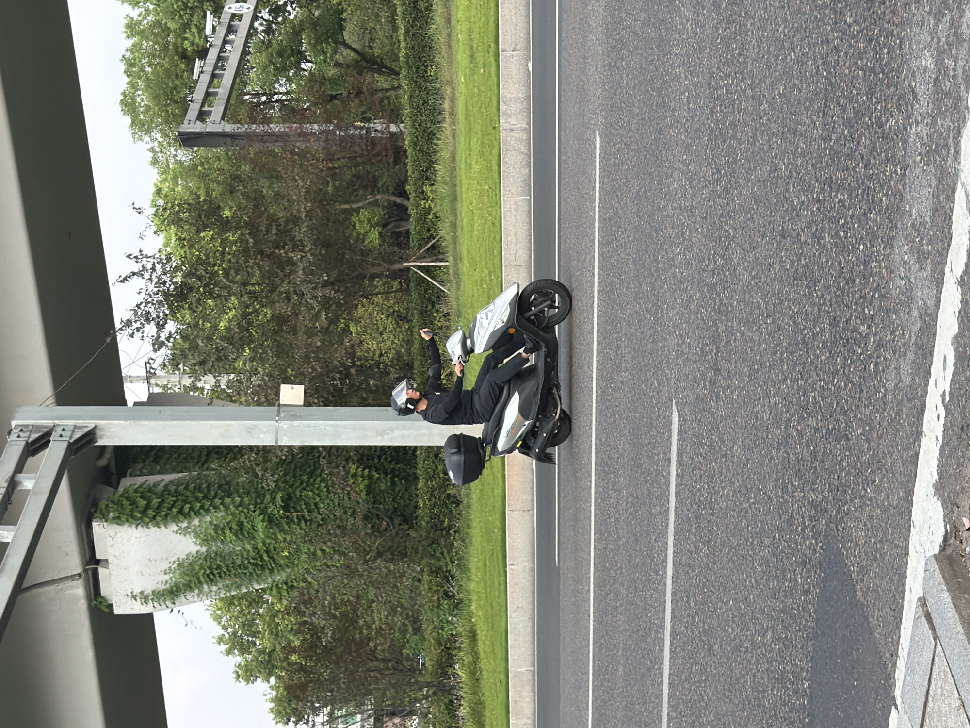 In the city of Jiangjin, China, a person is riding a silver and black scooter on a paved road. The rider is wearing a black outfit and a helmet. The scooter has a black storage box on the back. The road has white lane markings. To the left of the road, there is a grassy area with a concrete barrier. In the background, there is an overpass structure covered in green vines and trees. Further in the background, there are more trees and a traffic camera pole. The sky is overcast.