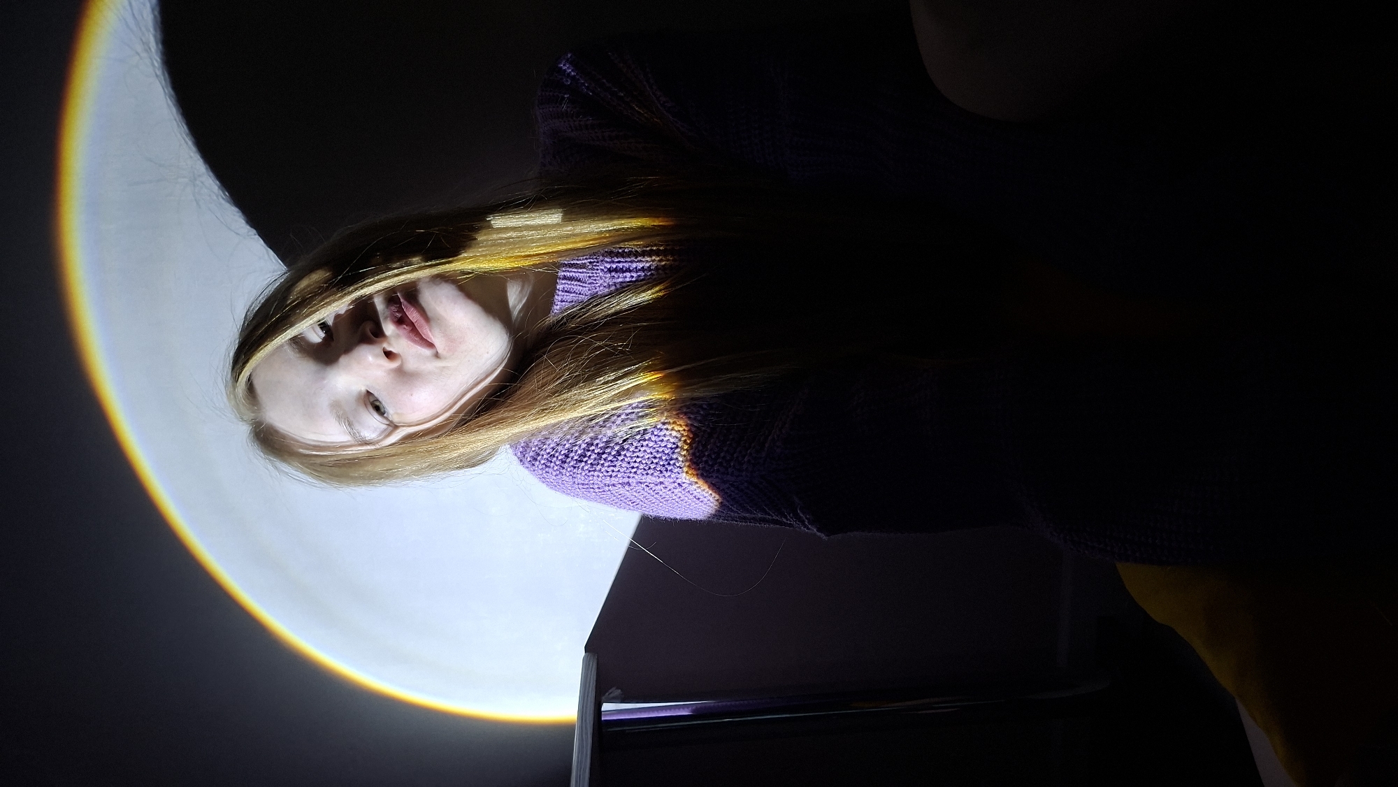 A pale-skinned person with light blonde hair is seen from a low angle in a dark room. The person is wearing a purple knitted sweater. A bright, multicolored arc of light (ranging from yellow to orange) is projected on the wall behind and to the left of the person, illuminating the left side of their face and neck. The person’s expression is neutral. Part of a dark object that is possibly a table can be seen below the person. The location is undetermined.