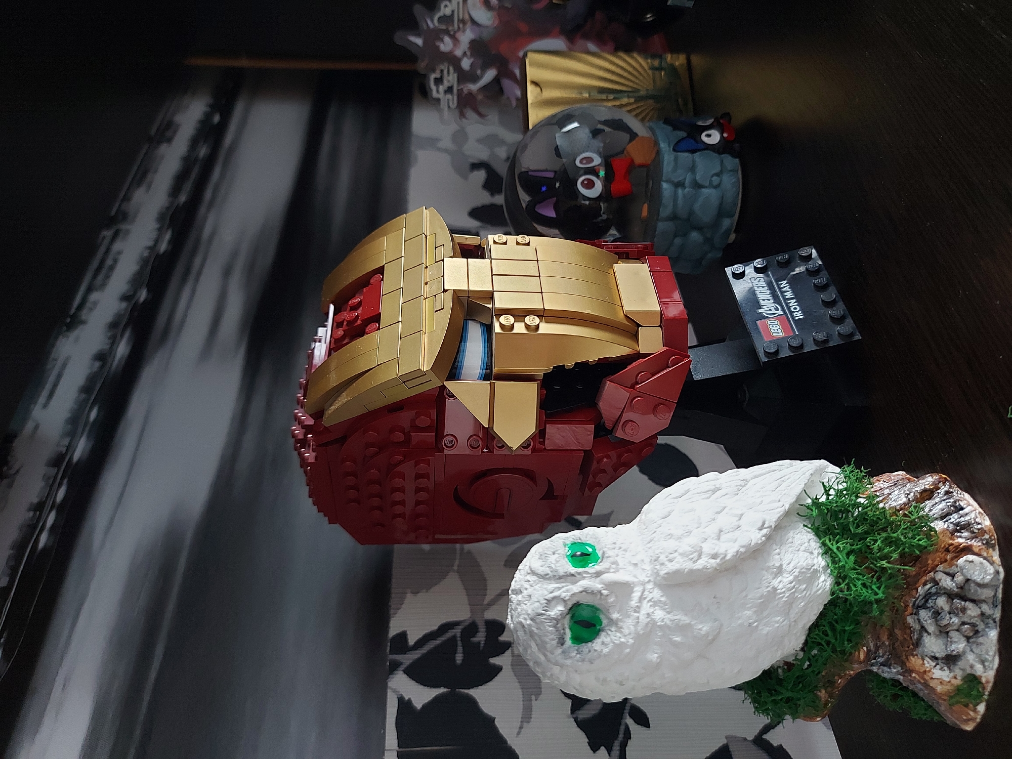 A slightly high-angle shot shows various objects on a surface. A Lego Iron Man helmet in red and gold dominates the frame. To the right, there is a small, white, sculpted bird with green eyes atop a base decorated with green moss and faux rocks. Behind the Iron Man helmet is a black Lego tile that says "LEGO AVENGERS IRON MAN." Further in the background are other items, including a spherical object with what appears to be a cat or rabbit figure inside, and what looks like a golden fan. The surface the objects are on has a gray and black print of what appears to be a waterfall. It is not possible to determine if this image was taken in Izhevsk, Russia. There are no people depicted in the image.