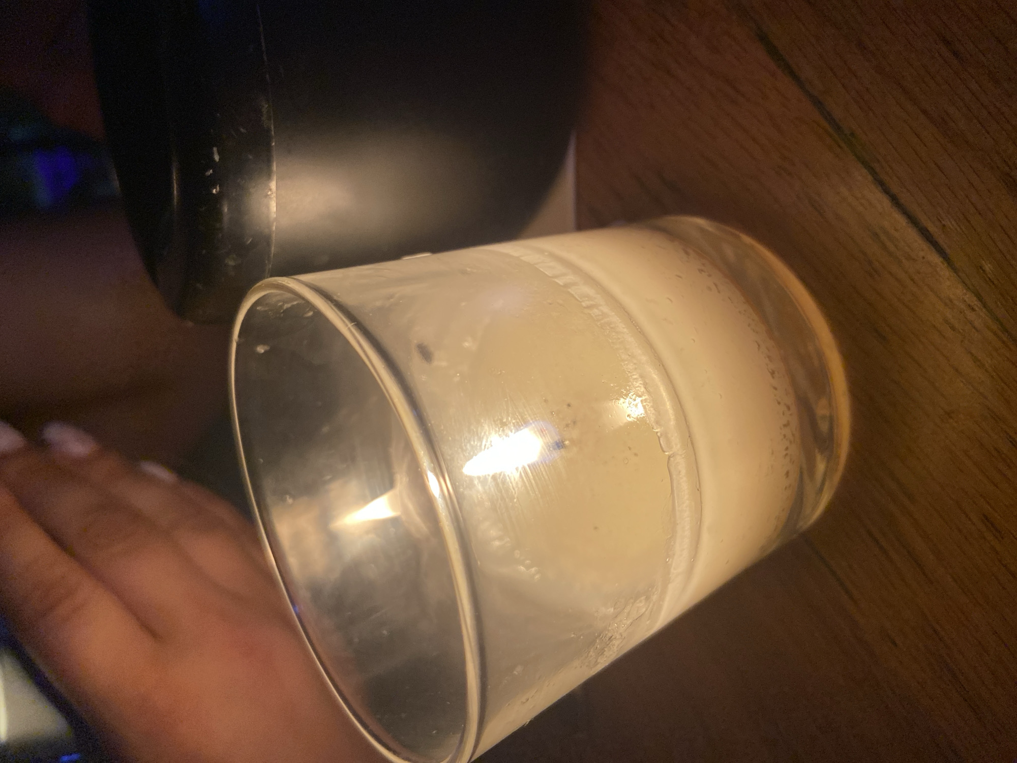 A close-up shot captures a lit candle in a glass holder, tilted slightly. The candle is a pale yellow, with a visible wick burning with a small flame. The glass holder is transparent and shows some condensation or residue on its interior surface. To the right of the candle, a dark, cylindrical object, possibly an ashtray or a container, is partially visible. In the upper left corner, a person's hand with manicured nails is in focus, suggesting proximity to the candle. The background is dark and out of focus, hinting at an indoor setting with low lighting. The overall impression is of an intimate or ambient atmosphere, perhaps in a restaurant or bar. The image does not contain information about the city of Sofia, Bulgaria.