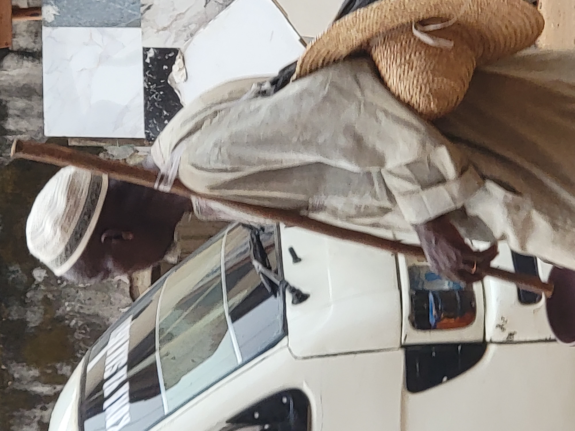A photograph, likely taken in Maiburiji, Nigeria, depicts a person standing alongside a light-colored, three-wheeled utility vehicle. The individual, appearing to be an adult male, has dark skin and is dressed in a light-colored, flowing garment, possibly a tunic or djellaba, paired with a white, patterned cap. A wide-brimmed straw hat rests on their back. With both hands, they hold a long, slender wooden stick diagonally across their body, possibly resting against the vehicle's front. Their head is slightly bowed, looking down towards the vehicle.
The vehicle itself is light-colored, resembling an auto-rickshaw or a small cargo trike, with a visible windshield and side mirrors. White text is discernable on the windshield, partially obscured and reversed, which appears to spell "PURE WATER," suggesting its function for transporting packaged water. The background features a wall composed of various square tiles, including plain white, grey marble-effect, and dark speckled designs. The setting appears to be outdoors during daylight hours, indicated by the natural lighting. The scene captures a moment of daily life, possibly a pause from work or travel, in an urban or semi-urban environment.