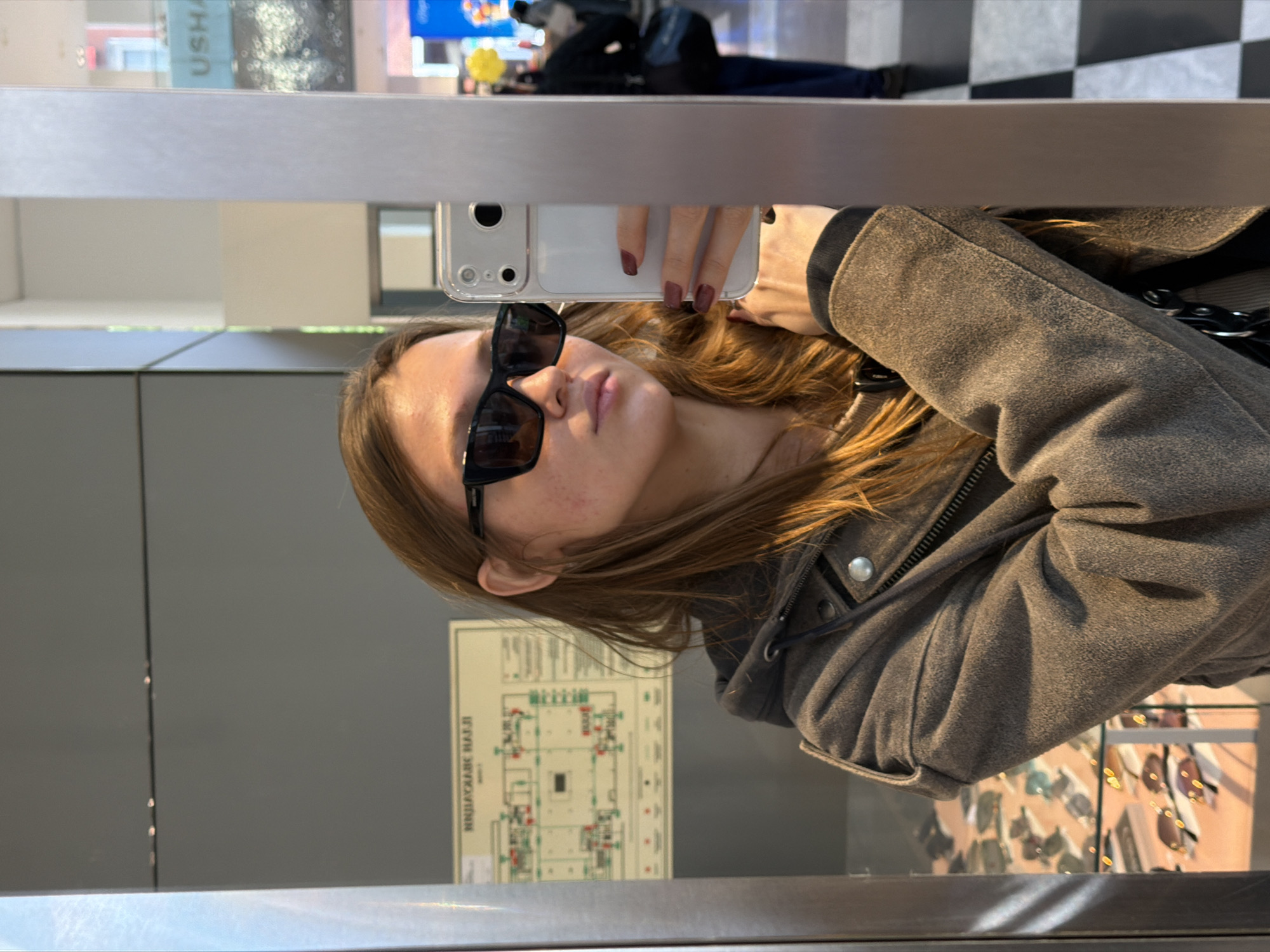 A woman is taking a selfie in a mirror. She is wearing sunglasses and a grey jacket. The reflection in the mirror shows a building evacuation plan in Russian (indicating "НИПКАУДЛПК НАДЖИ" - NIPKAUDLPK NADZHI), sunglasses on display, and other shoppers. Based on the text in Russian on the evacuation plan and general impression of the interior, it is plausible that the photo was taken in a commercial location in Moscow, Russia.