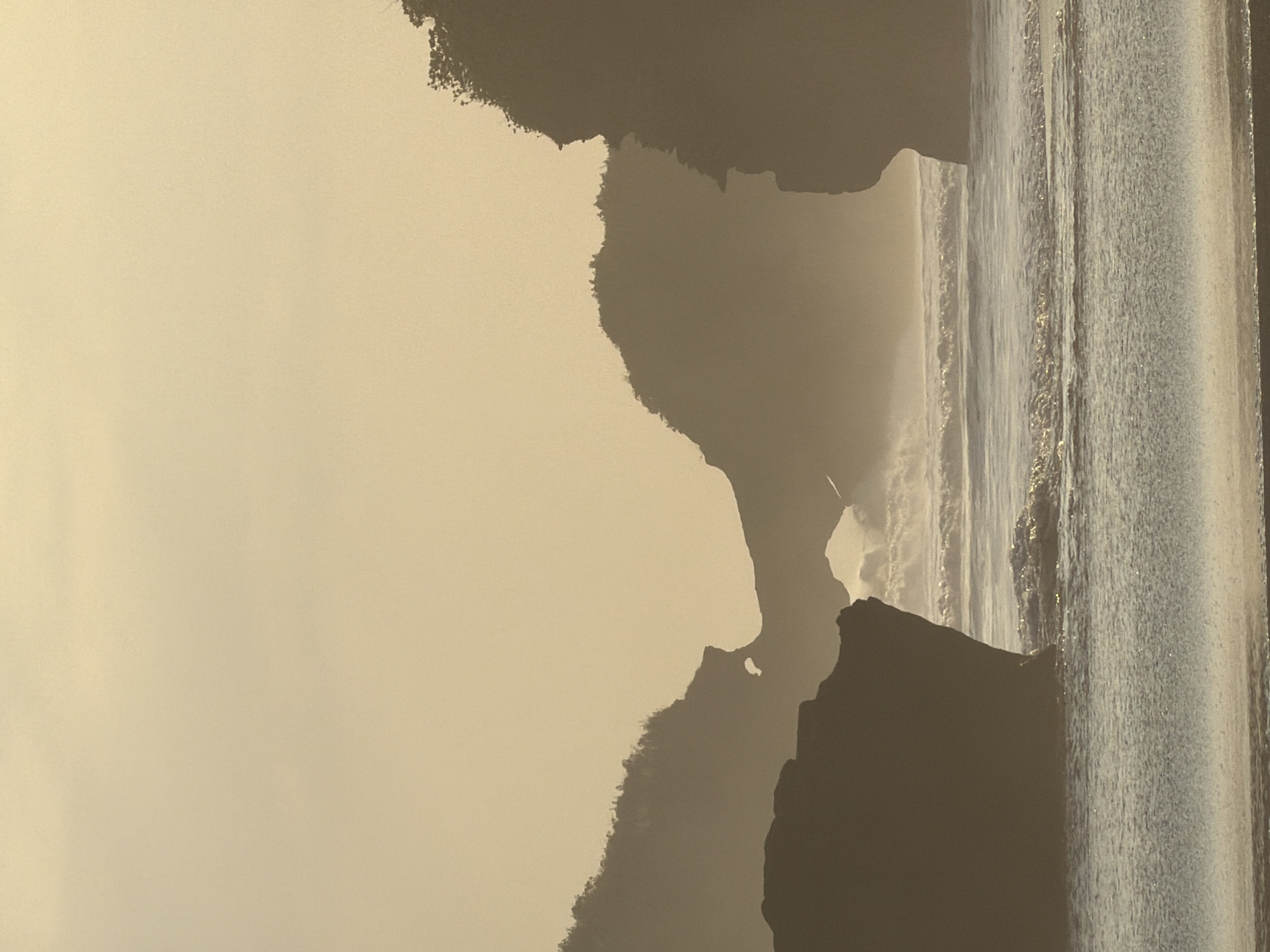 This vertical photograph captures a hazy coastal landscape in Selemadeg, Indonesia, at what appears to be sunrise or sunset, characterized by soft, diffused, warm-toned lighting. The upper two-thirds of the image consist of a uniform, light beige-yellow sky, free of distinct clouds, with atmospheric haze obscuring the distant horizon. The lower third features the ocean, with gentle waves breaking in the immediate foreground, transitioning to larger, cresting waves in the mid-distance that generate a visible mist.
Several prominent rock formations and cliffs are silhouetted against the diffused light. A large, dark, irregularly shaped rock occupies the lower-left quadrant, partially submerged in the water. In the mid-ground, a series of dark, undulating cliffs rise, topped with indistinct vegetation, and feature a distinctive natural arch or bridge formation through which light is visible. To the far right, a tall, dark cliff face extends into the frame from the edge, its upper profile also indicative of vegetation. The scene contains no discernible individuals, animals, or man-made objects.