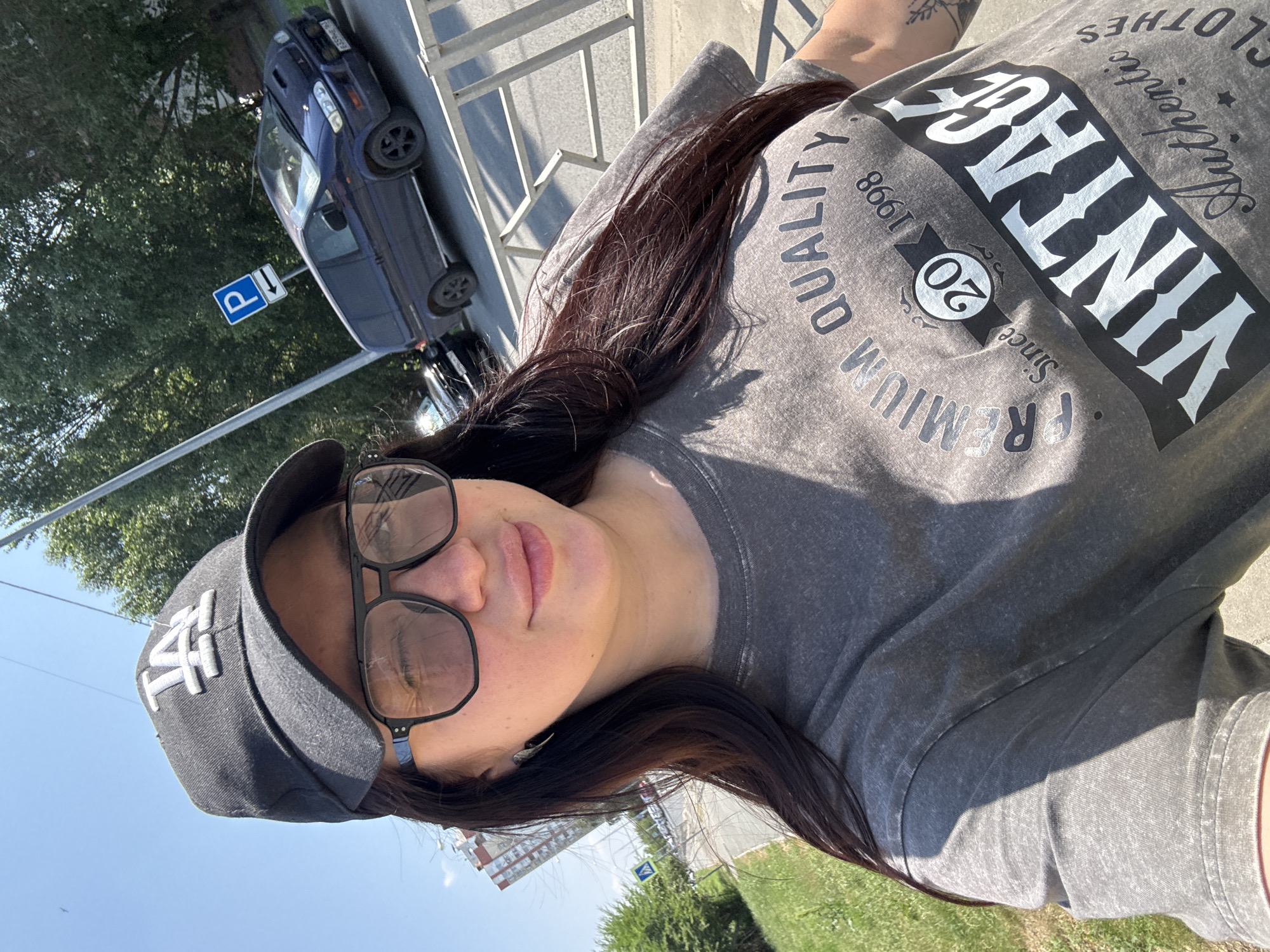 A young woman with long dark hair, wearing a grey t-shirt with a "VINTAGE AUTHENTIC CLOTHES SINCE 1998" graphic and a black baseball cap, takes a selfie outdoors on a sunny day. She is wearing large, dark-rimmed glasses and a winged earring. The background suggests an urban setting, possibly in Tolyatti, Russia, with a blue van parked on the street, a pedestrian crossing sign, a tall building in the distance, and trees lining the sidewalk. The woman is looking towards the camera with her left eye partially closed, and her lips are slightly parted. The overall impression is of a casual, everyday moment captured in an urban environment.