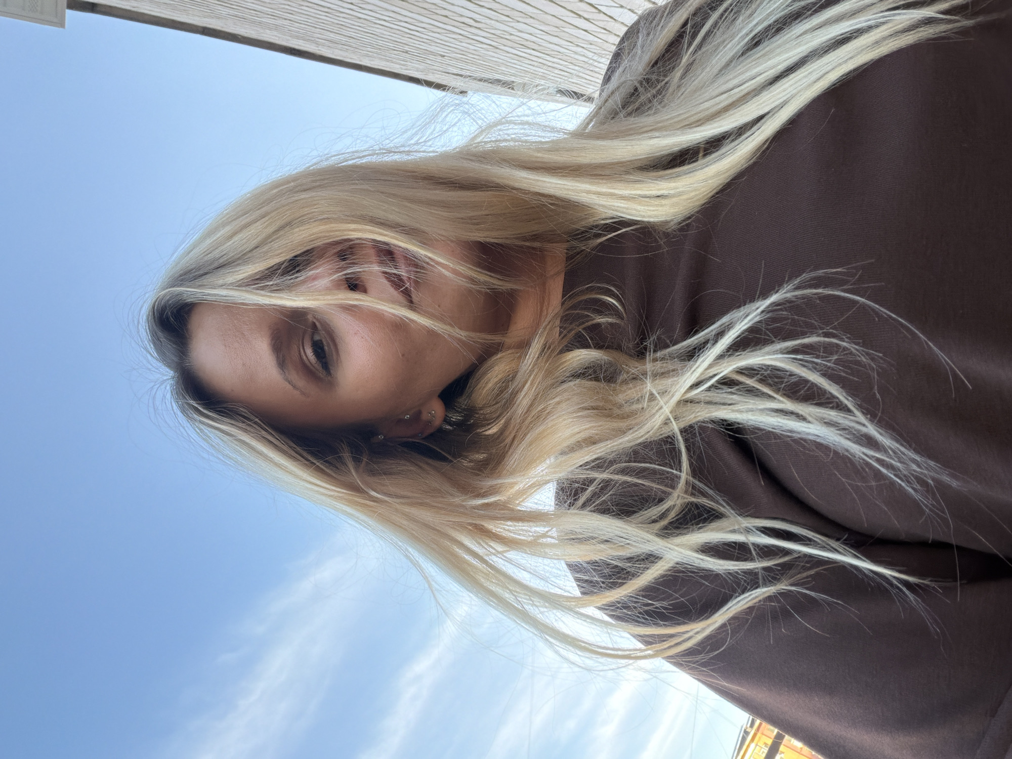 A woman with light blond hair and a brown shirt is looking down towards the camera. The sky is light blue with thin clouds in the background. A building is visible in the top left. The location is not discernible from the image.