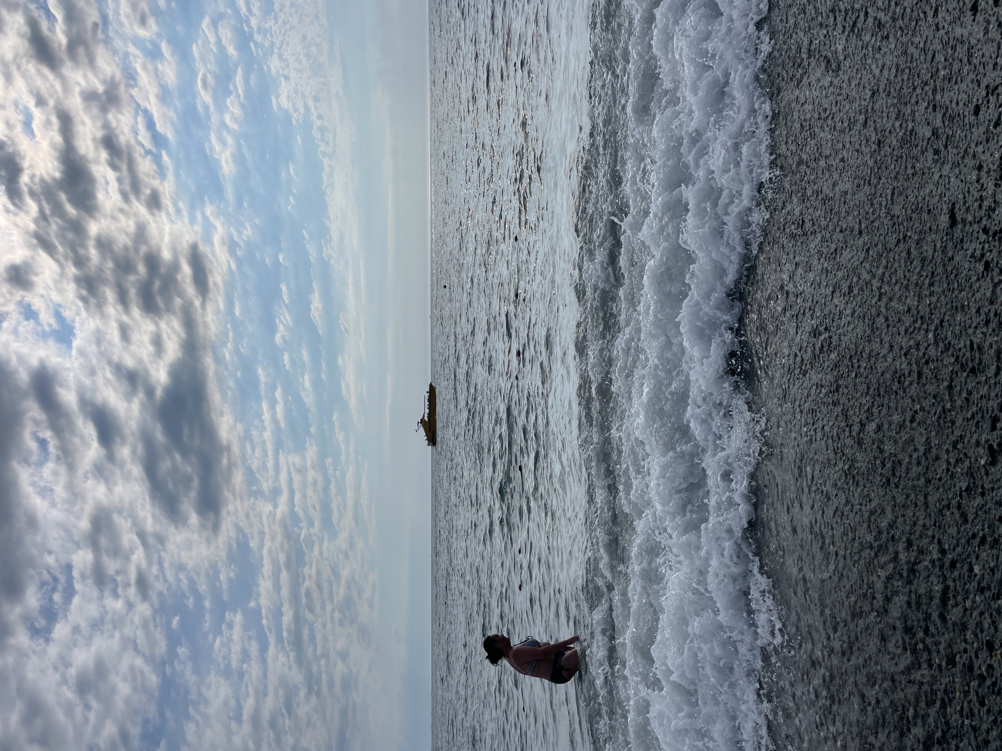 The image depicts a person in a bikini kneeling on a dark, pebbly beach as a wave breaks onto the shore. The water is a shimmering gray, reflecting the overcast sky. In the distance, a large yellow boat is visible on the horizon. The sky is filled with a mix of dramatic, dark clouds and lighter, wispy ones, suggesting a somewhat moody or transitional weather pattern. The overall setting appears to be a coastal area, with the texture of the beach and the vast expanse of the sea being prominent features. The time of day is likely daytime, given the ambient light, but the cloud cover prevents clear identification of direct sunlight. The image conveys a sense of peaceful contemplation or a moment of quiet enjoyment of the seaside. No visible text is present in the image. The location context of Sochi, Russia, aligns with the scene of a beach and the presence of a boat on the water.