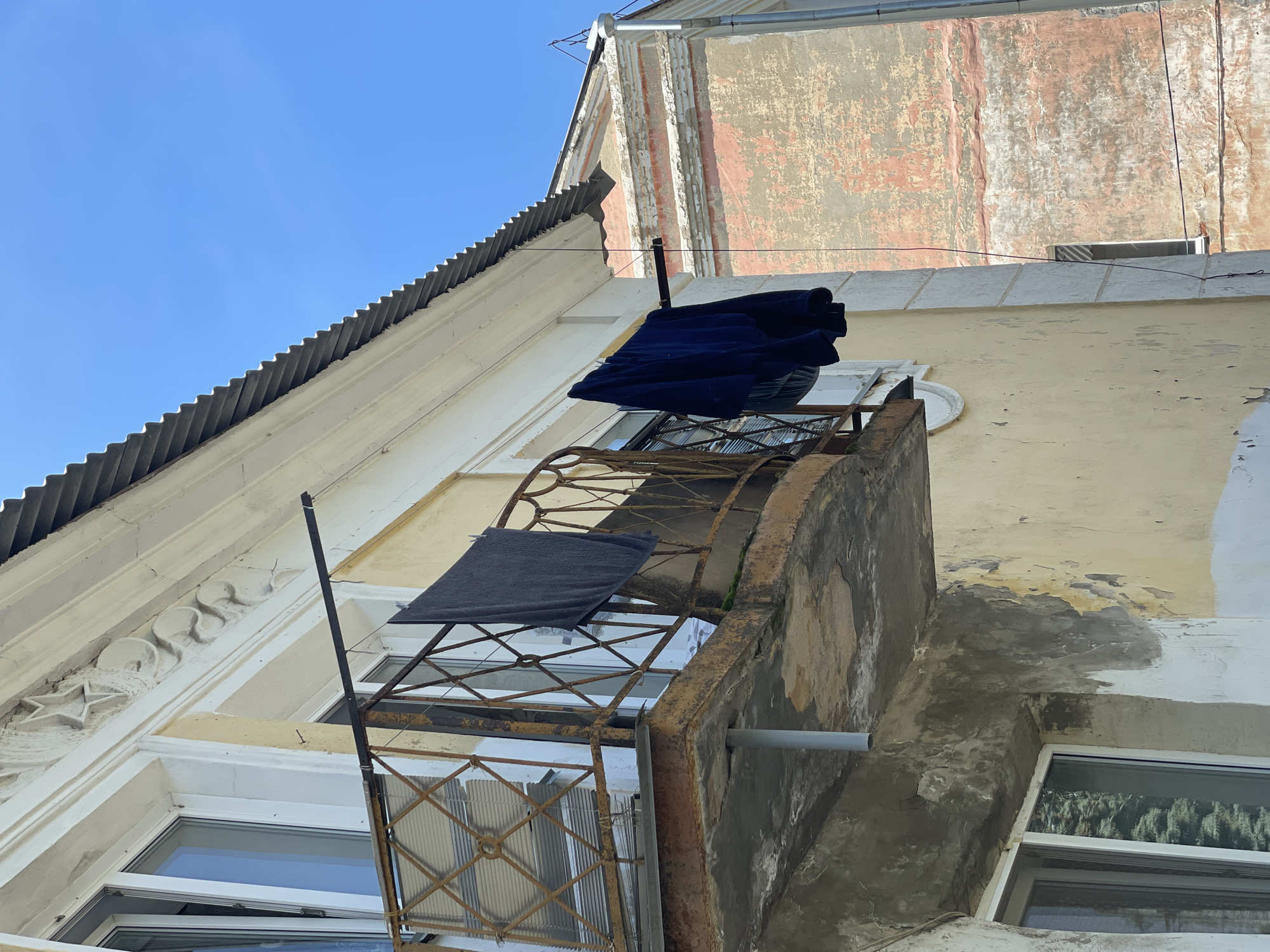 An exterior, low-angle, eye-level shot focuses on a weathered, yellow-painted building with a rusty balcony, against a clear blue sky. Laundry, consisting of a dark gray towel and a dark blue garment, hangs from a clothesline strung across the balcony railing. The balcony itself is constructed of rusted metal with a diamond pattern, and its concrete slab is chipped and peeling. To the right of the main building, another building with faded pink and orange plaster is visible. The architectural style suggests an older urban setting.