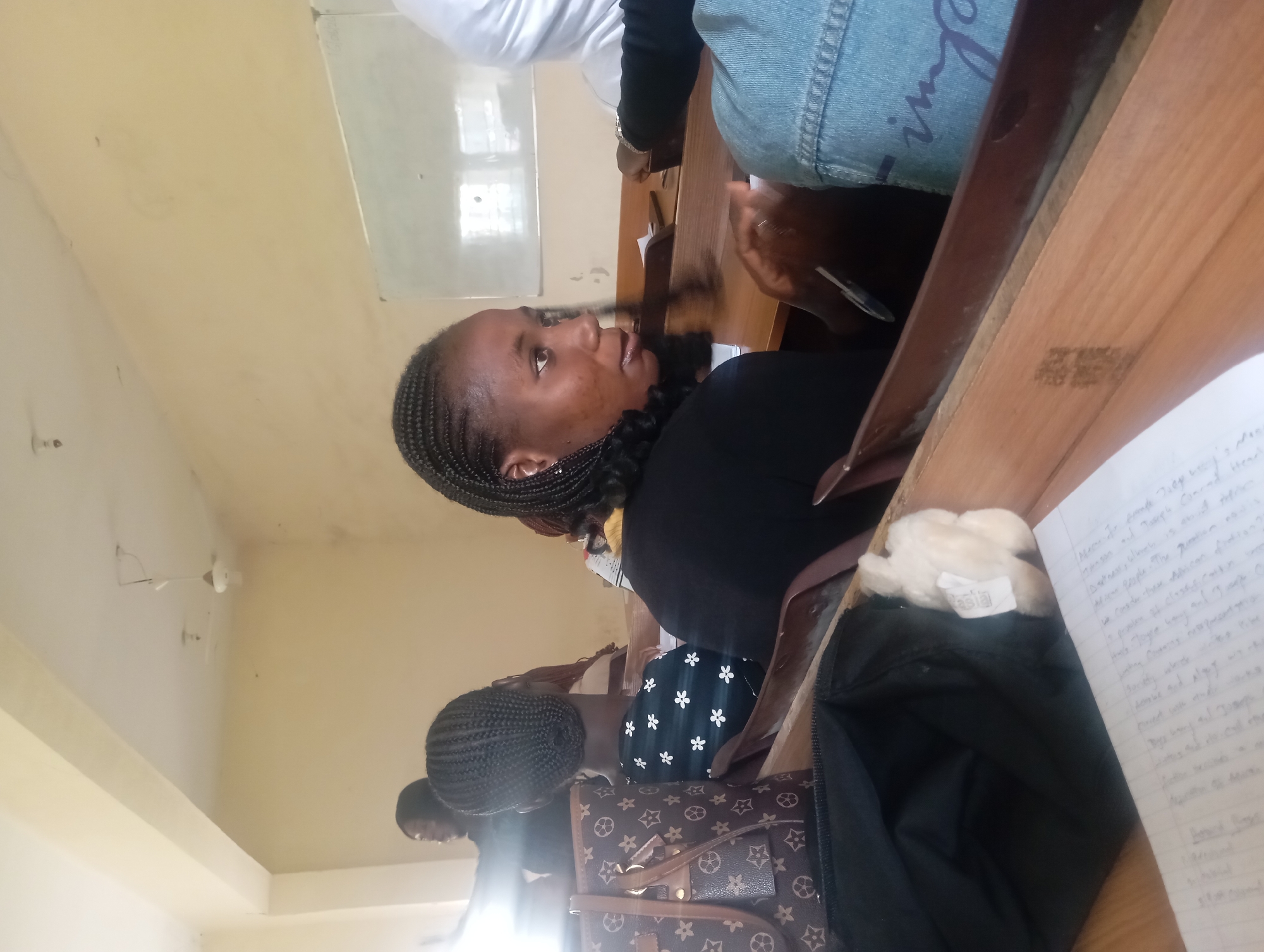 In an indoor classroom setting, several young women are seated at wooden desks, facing away from the camera. The room appears to be brightly lit, with a white board visible on the wall in the background. The students are dressed in casual attire, and some have their hair styled in braids. One woman in the foreground is looking to her left, while another is partially visible in the upper left corner of the frame, seemingly interacting with someone off-camera. A patterned handbag and an open black bag with some white material inside are placed on a desk in the foreground. The overall scene suggests a typical academic environment.