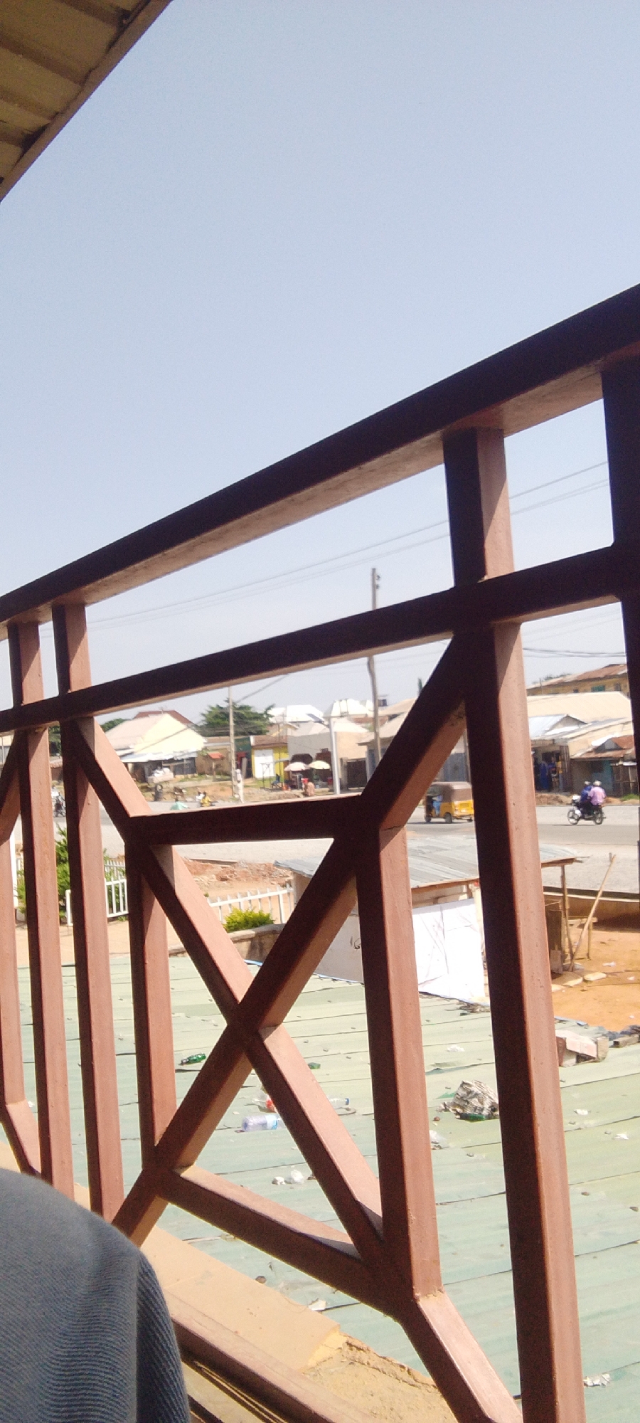 The image shows a street scene in Suleja, Nigeria, viewed from behind a brown wooden railing. The railing is angled, with vertical posts and horizontal beams forming a decorative pattern. In the background, buildings with light-colored roofs are visible, lining a paved road. Several people are present on the street. Some are walking, while others appear to be interacting near market stalls or small shops. A yellow auto-rickshaw and a motorcycle with two riders are on the road. The ground in the foreground, below the railing, appears to be a covered area with a light green surface, scattered with various pieces of litter, including plastic bottles. The sky is clear and light blue. The scene depicts a typical urban environment during daylight hours.