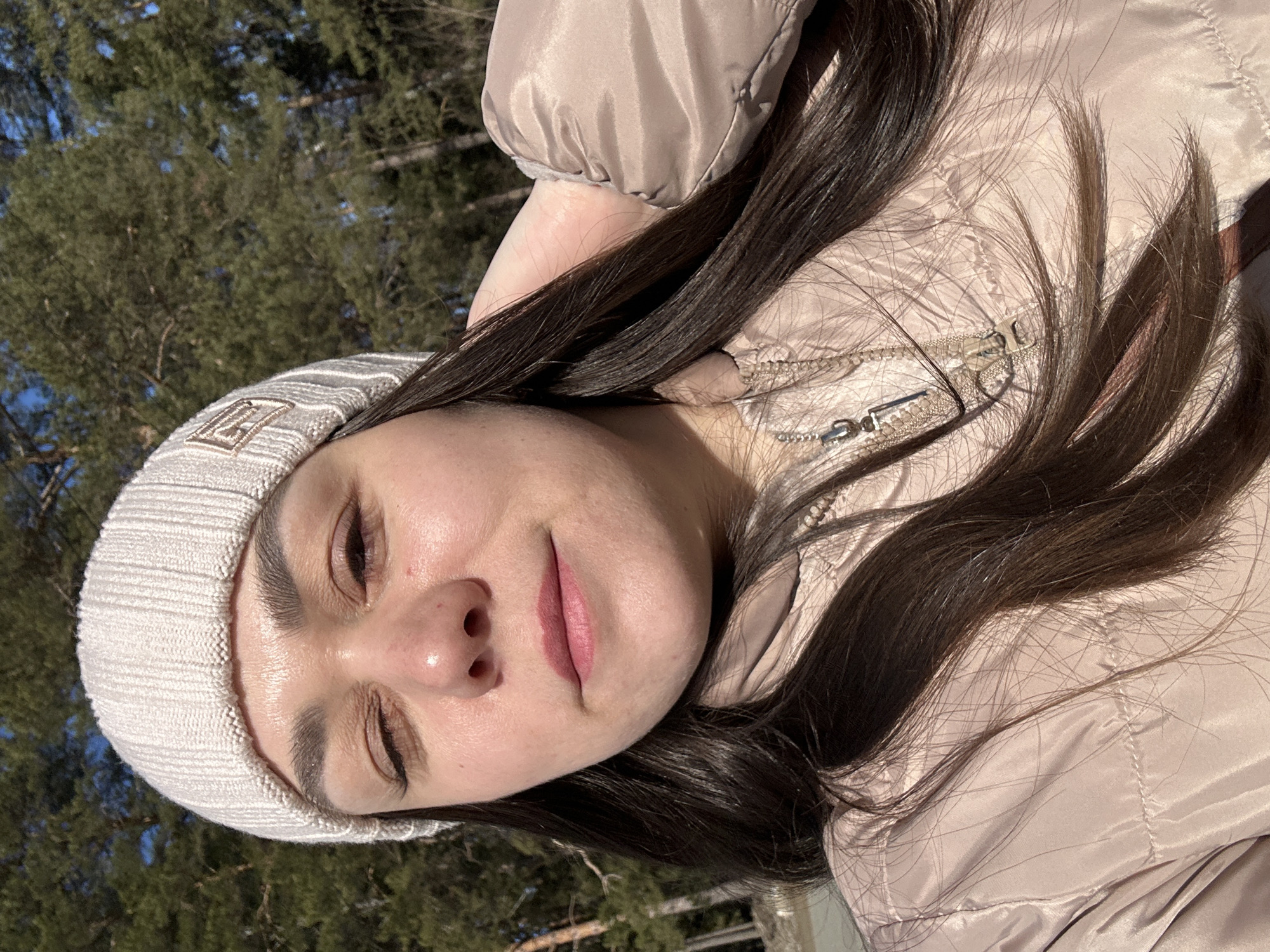 An adult female is depicted in a close-up outdoor portrait, facing direct sunlight. She has long, dark brown hair and is wearing a light beige ribbed knit beanie with a small, dark embroidered emblem on the front-right side. Her eyes are partially closed, consistent with squinting in bright light, and she has pink lipstick. Her right hand is raised and rests behind her head. She wears a light beige quilted puffer jacket with a visible zipper pull on the collar area. The background features dense evergreen trees under a clear blue sky. This natural, forested environment is consistent with outdoor settings found in or immediately surrounding the city of Chaikovsky, Russia.