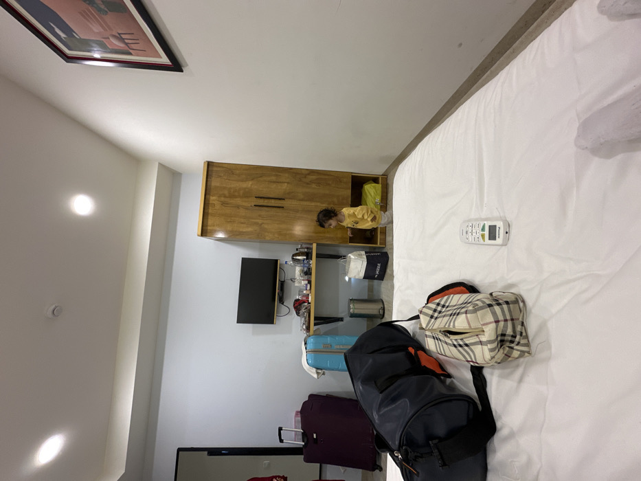 The scene is an indoor hotel room located in Vadodara, India. The perspective is low, looking towards a wall furnished with a desk, television, and wardrobe.
In the foreground, a large white bed occupies the lower half of the image. Resting on the white duvet are a dark blue duffel bag with orange accents, a beige plaid toiletry bag, and a white remote control. A white towel is partially visible on the far right edge of the bed.
In the midground, a light-skinned child, approximately 1-2 years old, with dark curly hair, wearing a yellow long-sleeved shirt and light-colored pants, stands near a tall wooden wardrobe on the right. The child is facing and looking into an open lower compartment of the wardrobe, which contains a folded yellow object. To the left of the wardrobe, a black flat-screen television is mounted above a wooden floating shelf or desk. On the shelf are multiple plastic water bottles, a black electric kettle, and other small items. Below the desk, a silver cylindrical trash can and two shopping bags, one white with "JADEUE" text and one yellow, are on the floor. Further left, a light blue wheeled suitcase is positioned against a light blue wall, next to a larger maroon wheeled suitcase.
The walls are predominantly light blue on the left and white on the right. A framed abstract artwork featuring pink and blue shapes is hung on the white wall. The white ceiling has multiple recessed circular spotlights and a perimeter light strip. A dark doorframe is partially visible on the far left. The floor appears to be a light-colored tile or wood laminate.