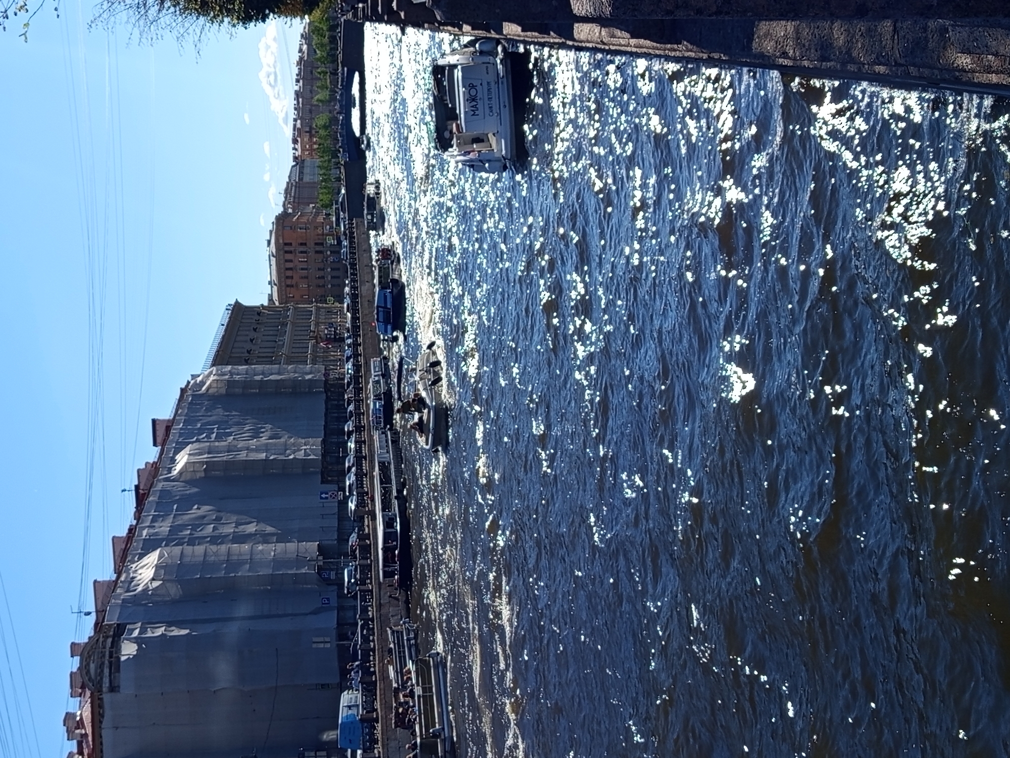 The image shows a wide canal in Saint Petersburg, Russia, with buildings lining the banks and several boats on the water. The sun is shining, creating numerous reflections on the choppy, dark blue water.
On the left bank, a large building is covered in scaffolding and grey tarps, suggesting ongoing renovation or construction. Several cars are parked along the embankment, and a blue van is visible. People are standing and walking along the sidewalk.
Several boats are navigating the canal. In the foreground, on the right side, a white motorboat with the word "МАЖОР" and "САНКТ-ПЕТЕРБУРГ" (Saint Petersburg) written on its stern is moving away from the viewer. Further up the canal, another motorboat carrying passengers is visible. Other boats, some appearing to be tour boats, are scattered throughout the waterway. A bridge crosses the canal in the distance, and more buildings are visible along the far bank. The sky is clear and blue.