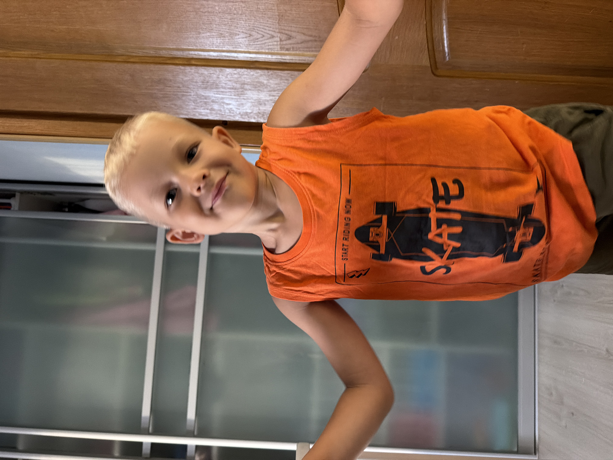 A young boy with blond hair and blue eyes is standing indoors, in front of a wooden door and a frosted glass closet. He is wearing an orange tank top with a black skateboard graphic and the words "START RIDING NOW" and "SKATE" printed on it. His arms are outstretched to the sides. The environment appears to be residential. The image does not provide any specific information that would allow for determination of its location within Sochi, Russia.