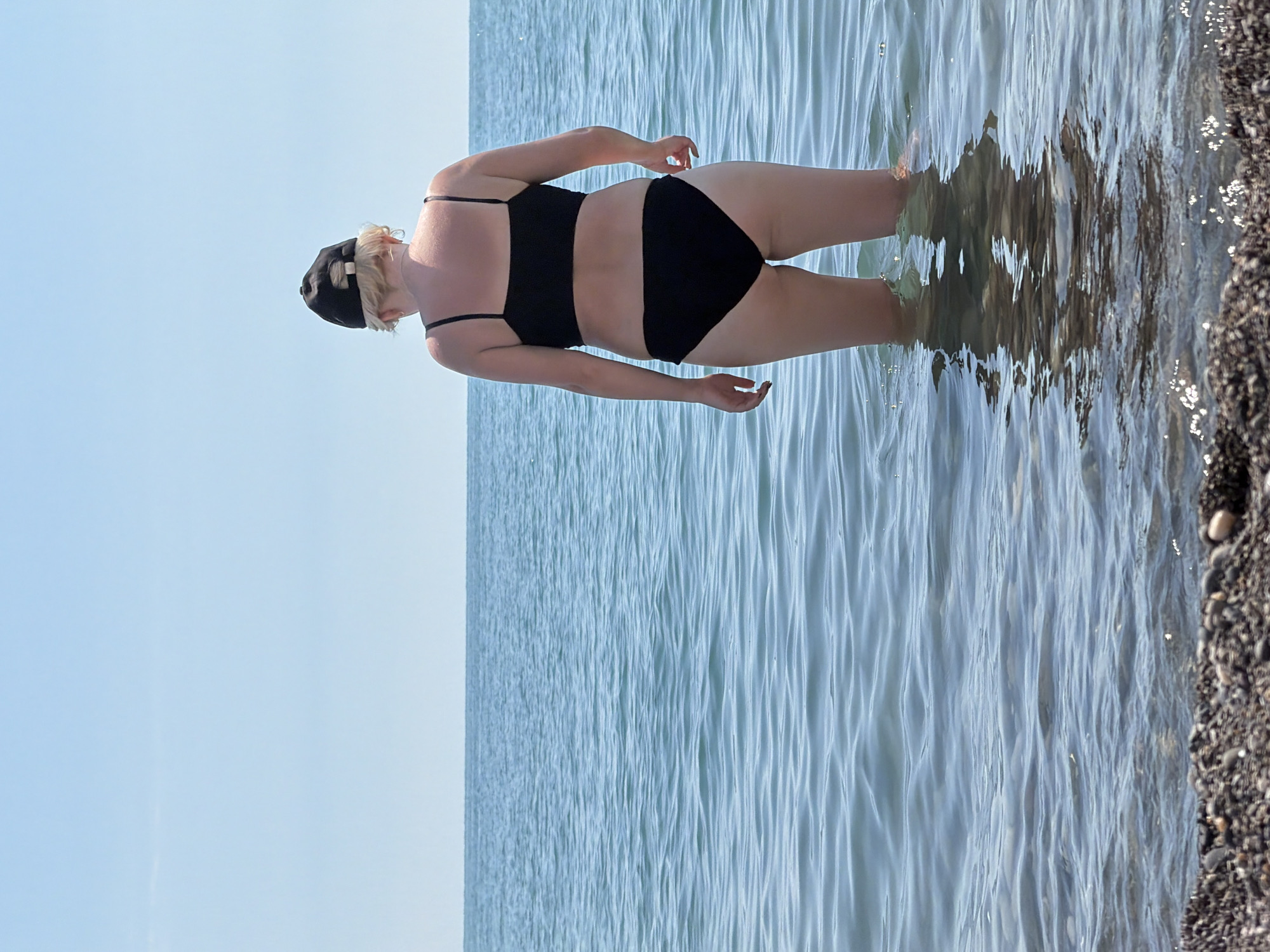 A person wearing a black bikini and a black baseball cap stands in the shallow water of the sea, facing away from the camera. The water is calm with small ripples, and the shoreline in the foreground is composed of small pebbles. The sky is clear and bright blue, with the horizon where the sky meets the sea visible in the background. This scene is depicted at a beach in Sochi, Russia.