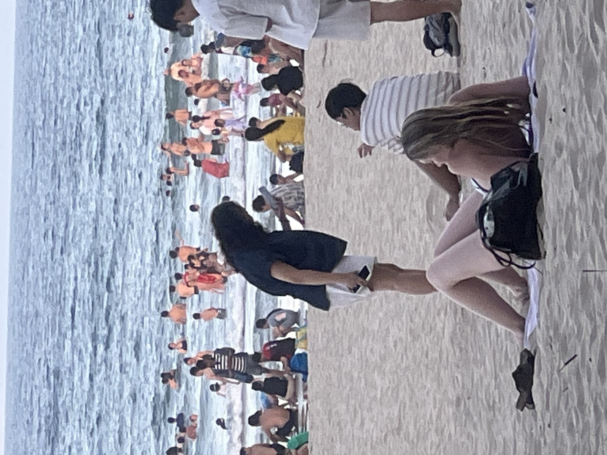 The image depicts a crowded beach scene at My Khe Beach in Da Nang, Vietnam. In the foreground, a woman with long blonde hair is lying on a towel on the sand, facing to her left and looking away from the camera. She is wearing a bikini. Next to her, a person wearing a striped shirt is sitting up on the sand, facing away from the camera and looking to their left. Further back on the beach, a person with dark, long hair is standing and holding a phone. They are wearing a dark top and white shorts. Behind them, numerous people are gathered in the water, some swimming and others standing. The sea is choppy with moderate waves. The beach is sandy and appears to be densely populated with beachgoers. In the background, the horizon shows a hazy sky. There are no prominent landmarks visible in this particular frame to definitively identify the specific location within Da Nang, but the overall scene is characteristic of a popular tourist beach.