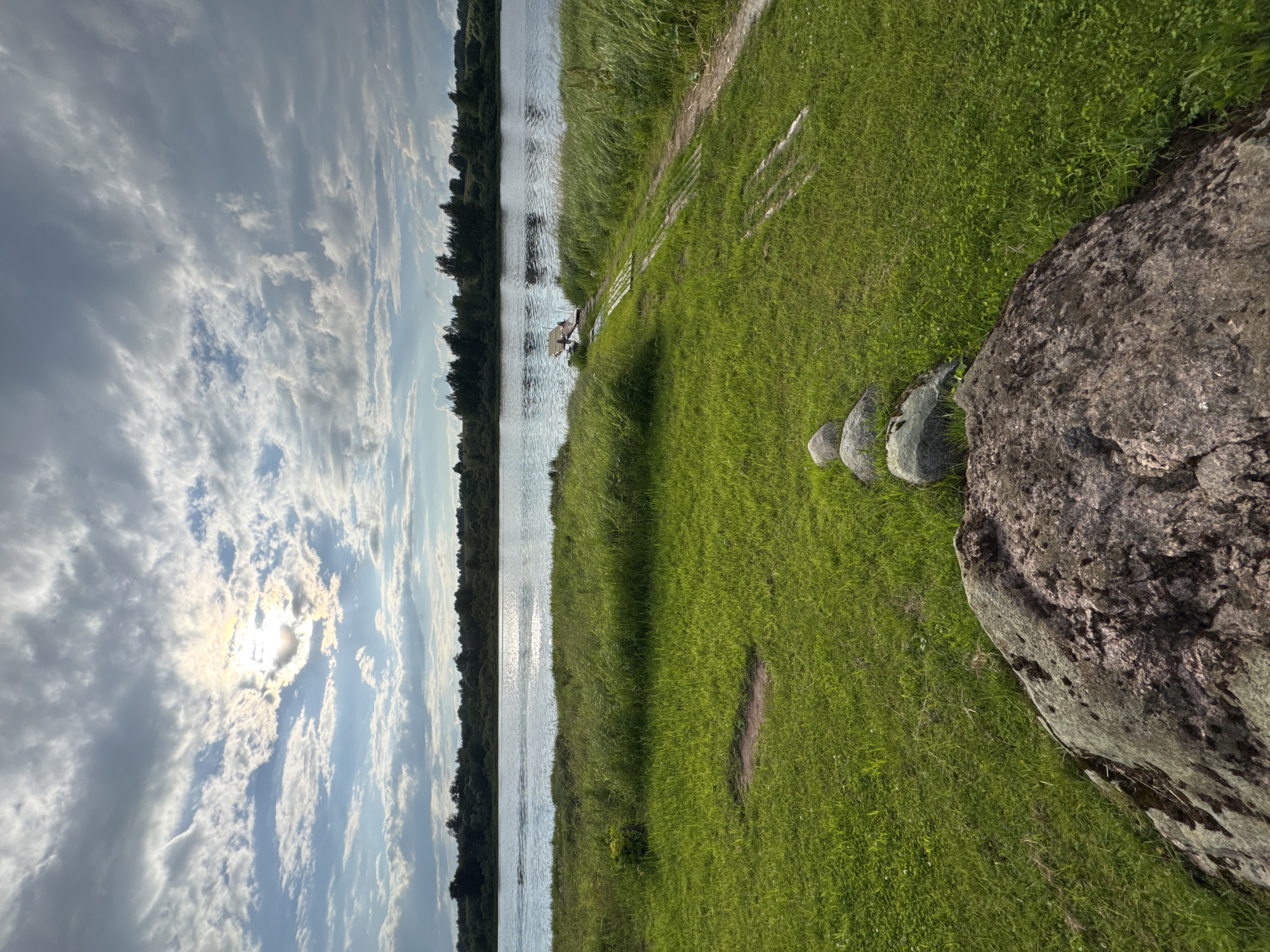 This is a wide-angle, landscape photograph taken at ground level, looking across a grassy embankment towards a wide, calm river under a dramatic, cloudy sky. The sun is partially obscured by thick, wispy clouds, casting a bright, diffused light across the scene, with rays of sunlight visible. In the foreground, a large, rough-textured boulder sits prominently, with several smaller, rounded stones arranged in a line behind it, leading towards the river. The grass is lush and green, with some patches of shorter, more manicured lawn and areas of taller, wilder grass near the water's edge. A narrow dirt path with wooden planks running alongside it, possibly a makeshift ramp, leads down to a small wooden dock jutting out into the river. Across the river, a dense line of dark green trees forms the horizon, with a few sparse trees visible in the distance. The water reflects the bright sky, creating a shimmering, silvery surface. The overall impression is one of a peaceful, natural landscape with a hint of human intervention. The location is described as Sitno, Russia.