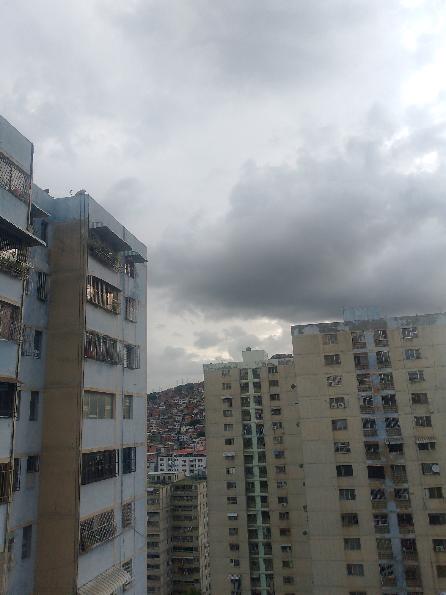 This image captures a view of the Petare neighborhood in Caracas, Venezuela, under a heavy, overcast sky. The foreground is dominated by two large, multi-story apartment buildings. The building on the left is painted a pale blue with concrete accents and features numerous balconies, windows with metal bars, and awnings. The building on the right is a lighter beige color, also with many windows and balconies, some of which appear damaged or weathered.
In the background, the dense, informal housing that characterizes Petare climbs the hillsides, a sprawl of red-tiled roofs and colorful walls. Several other apartment buildings, including some appearing taller and more modern than those in the foreground, are visible in the mid-ground, interspersed with the hillside development. Electrical power lines are faintly visible against the sky behind some of the buildings. The overall atmosphere is somber, with the dark clouds suggesting impending rain. There are no people or discernible actions visible in the frame.