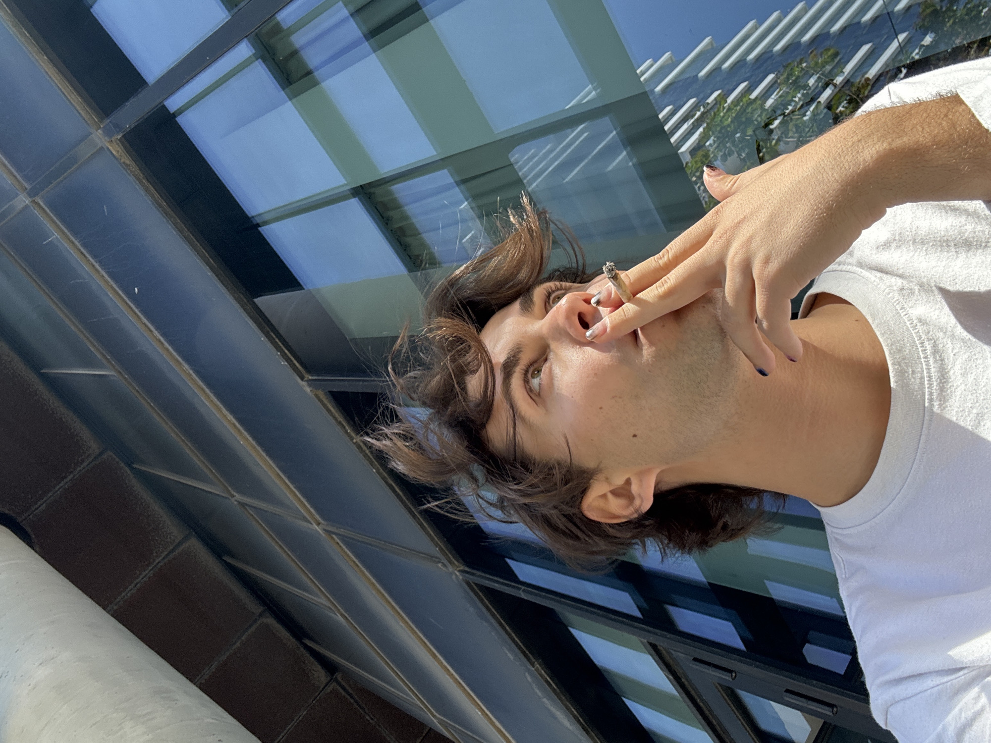 A person with brown hair and a white t-shirt is holding a cigarette in front of a modern building with glass windows and metal accents. The windows reflect the sky and nearby trees. The image is taken from a low angle, looking up at the person and the building. The person has painted fingernails. It is not possible to determine the location of this image in the city of Tel-Aviv.