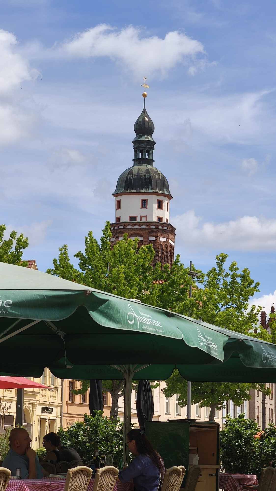 The image shows a tall, multi-tiered tower topped with a black dome and a gold cross, likely part of a church or historical building. The tower's lower sections are constructed of red brick, while the upper tiers are white with red-framed windows. Lush green trees partially obscure the tower. In the foreground, a green umbrella with the word "Amarone" printed on it provides shade to outdoor seating at a cafe. Several people are seated at the cafe, including a man wearing a blue shirt on the left and a woman with long brown hair wearing a blue shirt on the right. The tables are covered with red and white checkered tablecloths. Buildings line the street in the background, creating an urban setting, city of Unknown, Unknown. The sky above the tower is blue with scattered white clouds.