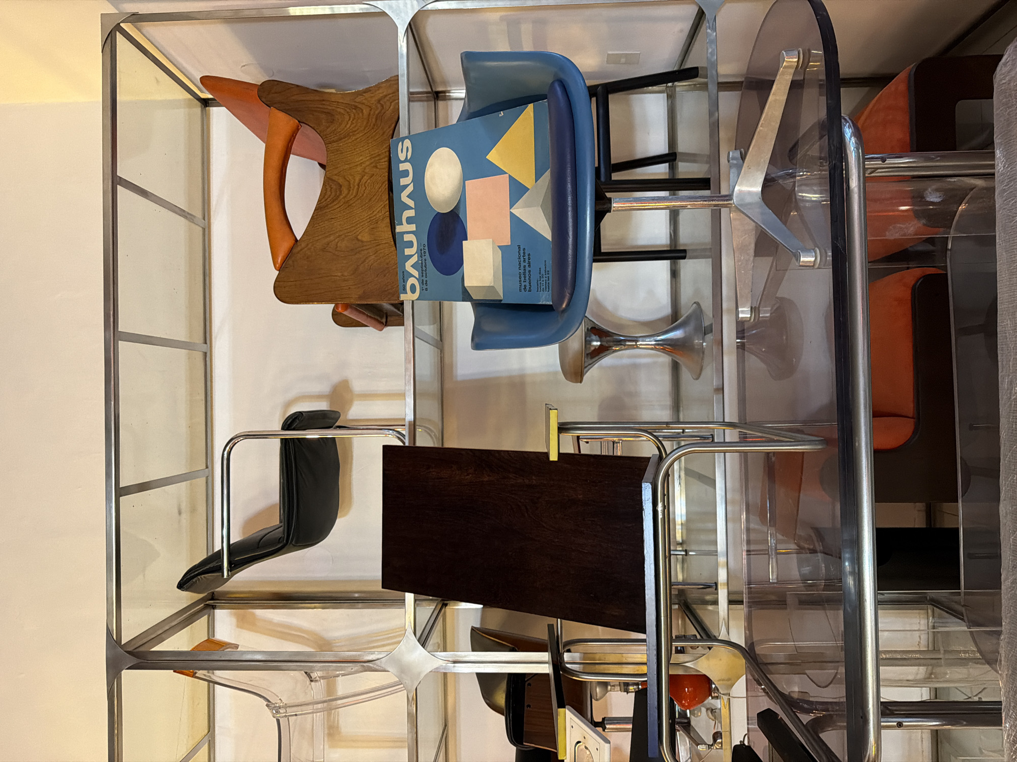 The image displays a collection of chairs and other furniture items, seemingly arranged within a shelving unit with a metal frame and transparent shelves. The furniture includes a wood-and-orange seat, a blue chair holding a "Bauhaus" pamphlet referring to an exhibition at the Museo Nacional de Buenos Aires, a black seat with chrome frame, a wooden table top, an orange chair, a transparent chair, and other furniture of mixed styles. The setting appears to be an interior space, possibly a showroom or storage area. The location in the city of Buenos Aires, Argentina is indirectly indicated by the pamphlet on the blue chair. The image appears to be a casually-composed shot and lacks any human presence or interaction.