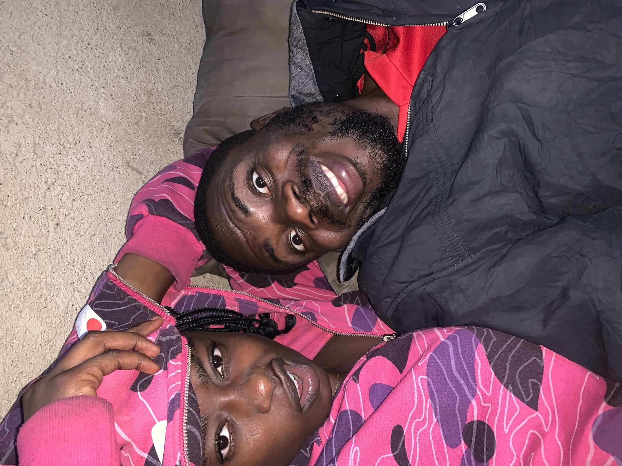 This is a selfie of two people, a woman and a man, lying down, possibly in a domestic setting. The woman is in the foreground on the left, wearing a pink and purple camouflage hoodie. She has dark skin, dark eyes, and her hair is in braids. Her mouth is slightly open, and she appears to be looking at the camera. The man is to her right, also looking at the camera with wide eyes and a large smile. He has dark skin and a beard, and is wearing a black jacket over a red shirt. The background appears to be a textured wall and a brown surface, possibly a couch or bed. The lighting is dim, suggesting it might be indoors at night. There are no specific landmarks or indicators that would place this image in Rafin Par, Nigeria; it's a generic indoor scene.
