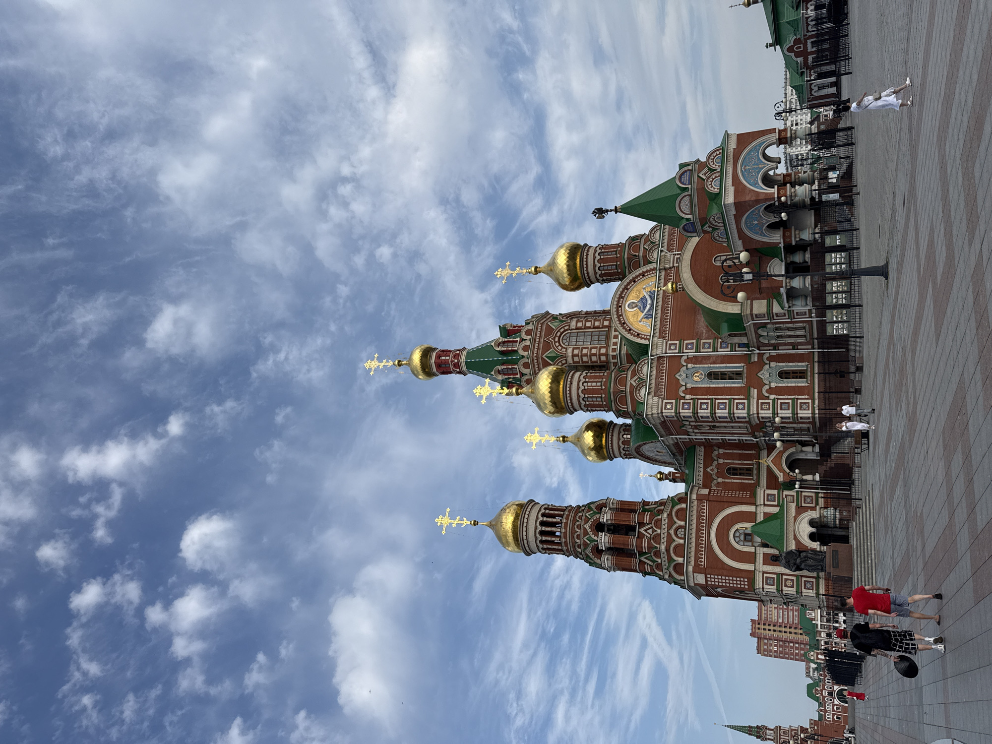 The image depicts a view of the Cathedral of the Annunciation in Yoshkar-Ola, Russia, under a cloudy blue sky. The cathedral is a large, ornate structure with red brick walls, green roofs, and multiple golden domes topped with crosses. In the foreground, a paved plaza is visible, with several people walking. Two men are walking away from the camera, one carrying a black circular object. To the right, a woman in a white dress is walking towards the cathedral. In the background, other buildings, including a tall tower and a modern residential building, can be seen. The scene appears to be a public square in front of the religious landmark.