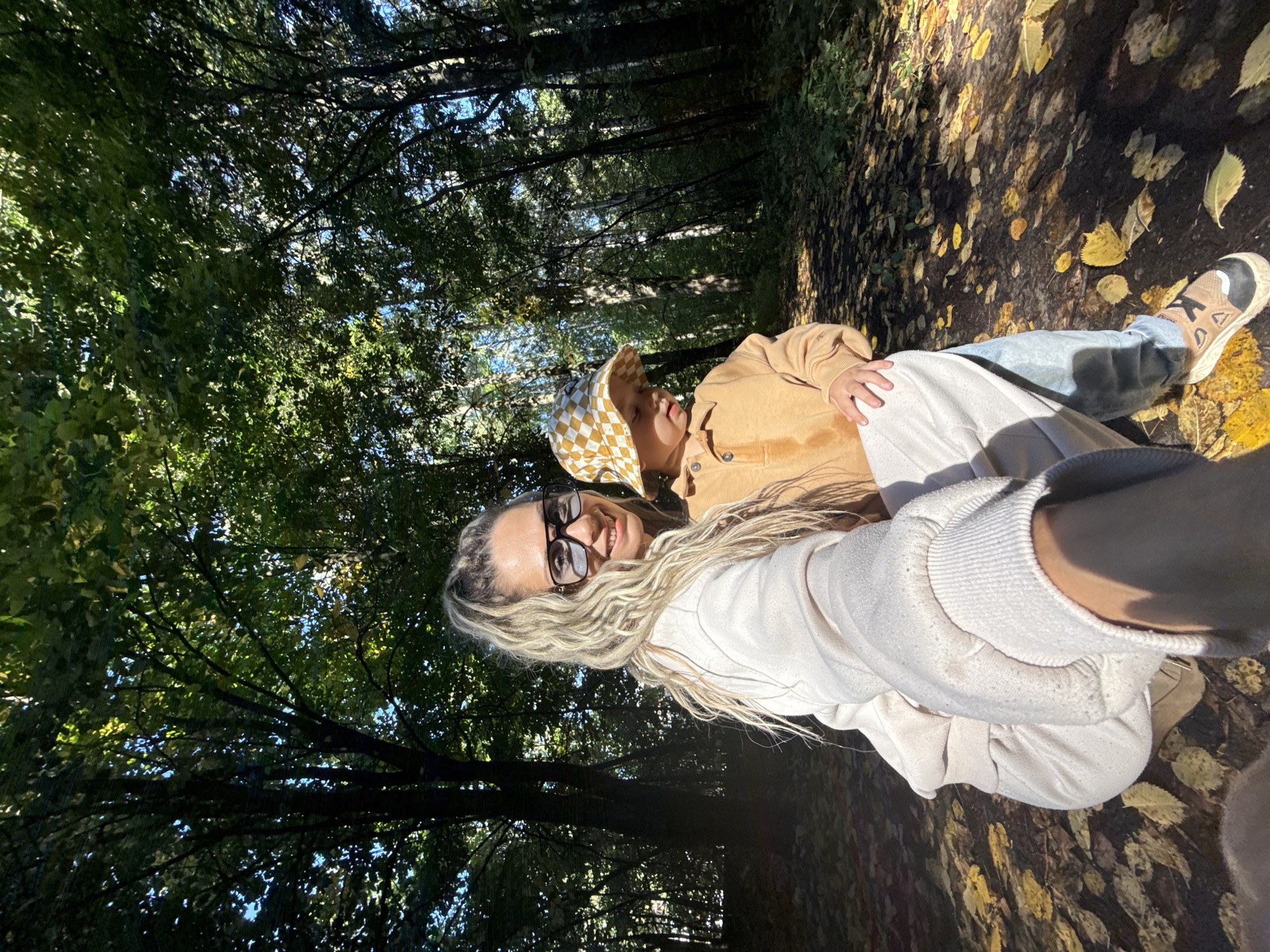 In an outdoor setting resembling a forest path, a woman with blonde, curly hair pulled into a high ponytail and wearing oversized sunglasses and a light-colored sweatshirt takes a selfie. Beside her, a young child wearing a checkered bucket hat and an orange sweatshirt looks down. The ground is covered with fallen yellow and brown leaves, indicating an autumn season. The background consists of dense trees with lush green foliage, dappled with sunlight. The overall scene suggests a casual outing in a wooded area, possibly in or near Rusanovo, Russia, given the context.