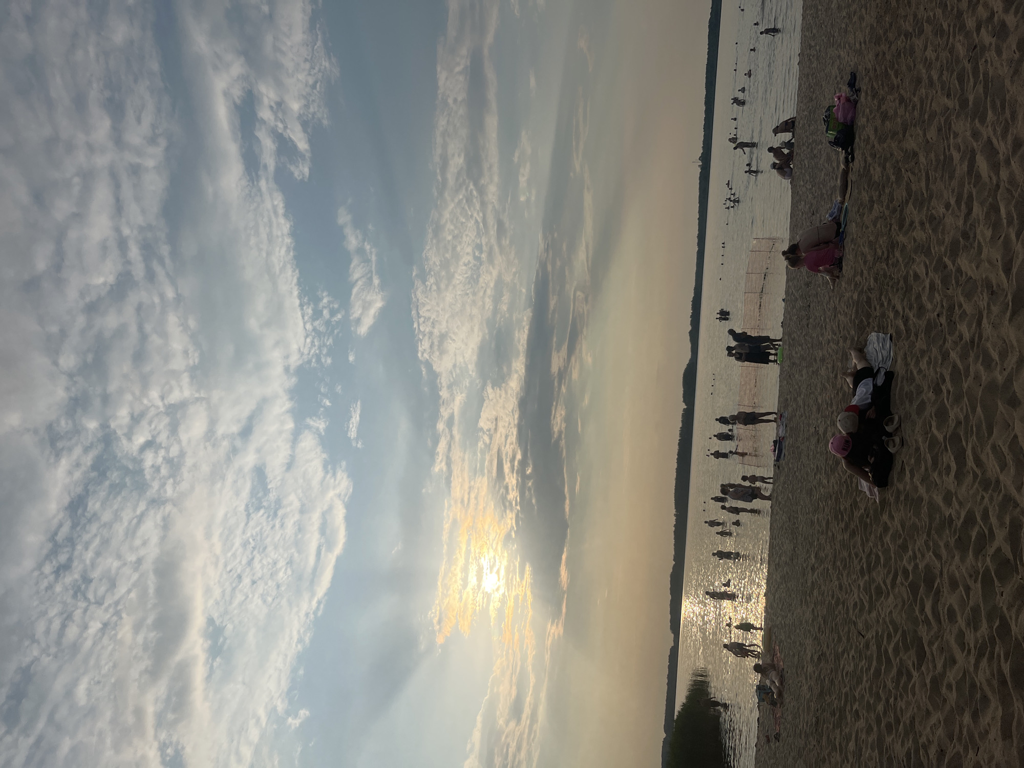 The image captures a scene at a sandy beach with people enjoying the water and the shoreline. The sky is filled with a mix of bright clouds and streaks of sunlight, suggesting it is either sunrise or sunset. The sun's rays are prominently visible, shining through the clouds and reflecting on the water.
Several individuals are visible in the water, some appearing to be swimming or wading. On the sandy beach, other people are seen relaxing, sitting, or lying down. Some are engaged in conversation or simply enjoying the beach atmosphere. The sand is textured with footprints and ripples, indicative of a well-used beach.
The overall mood appears to be peaceful and recreational, with people spending a pleasant day outdoors. The water is calm, and the distant shoreline is lined with trees, providing a natural backdrop. The specific location is identified as Загародскі сельскі Савет, Belarus. There is no visible text in the image.