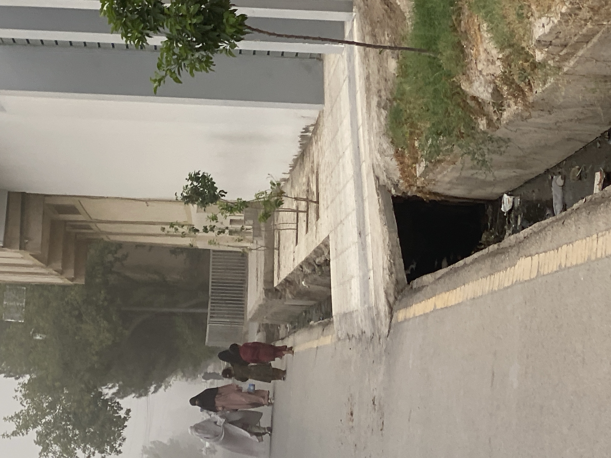 In Kandahar, Afghanistan, two women in full veils, one in a pale grey and the other in a black burqa, walk on a paved street. They are followed by two young children, one in a dark green garment and the other in a maroon outfit, also walking on the street. Adjacent to the street is a sidewalk with young trees planted along it. A drainage ditch runs alongside the sidewalk, containing dark, stagnant water and visible litter. Behind the sidewalk and trees, a white and grey building stands. The scene is characterized by a hazy, possibly dusty, atmosphere.