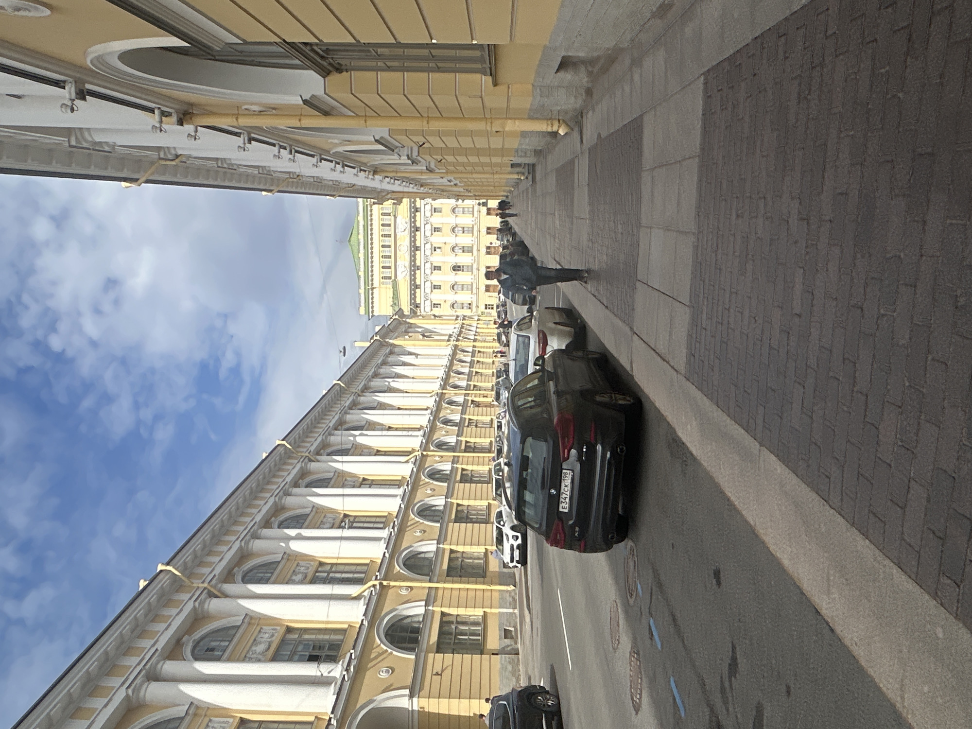 A low-angle, vertical, eye-level shot captures a street scene in a European city under a partly cloudy sky. On the left, a grand, yellow building with white columns and arched windows stretches into the distance. Parked cars, including a black SUV in the foreground, line the street. A man in a dark jacket walks away from the camera on the right side of the frame, along a sidewalk paved with dark bricks. The sidewalk is bordered by another yellow building, similar in style to the one on the left. In the background, a large, ornate yellow building with a green roof is visible. The scene appears to be a quiet daytime street with a few pedestrians and vehicles.