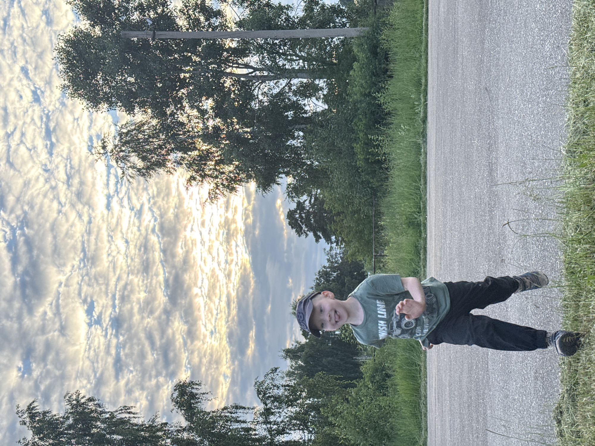 A young boy wearing a green t-shirt, black pants, a baseball cap, and athletic shoes stands on the side of a paved road. He is looking towards the camera with his tongue sticking out. The t-shirt has a graphic design and the words "FINISH LINE" and "OUR ADVENTURE" visible. The setting is outdoors, with trees and lush green grass on either side of the road. The sky is filled with dramatic, textured clouds, illuminated by what appears to be the setting sun, suggesting it is late afternoon or early evening. The overall mood is playful and candid.