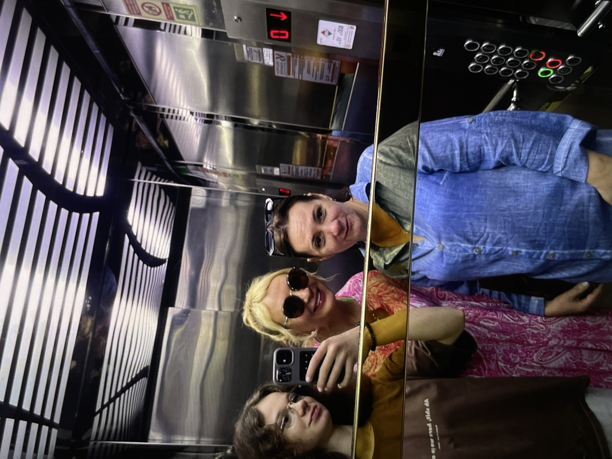 Three individuals are captured in a mirror reflection inside an elevator. The elevator panel indicates they are on the ground floor "0" and has an array of buttons with green and red indicators. The interior of the elevator appears modern with metallic surfaces and vertical slat details.