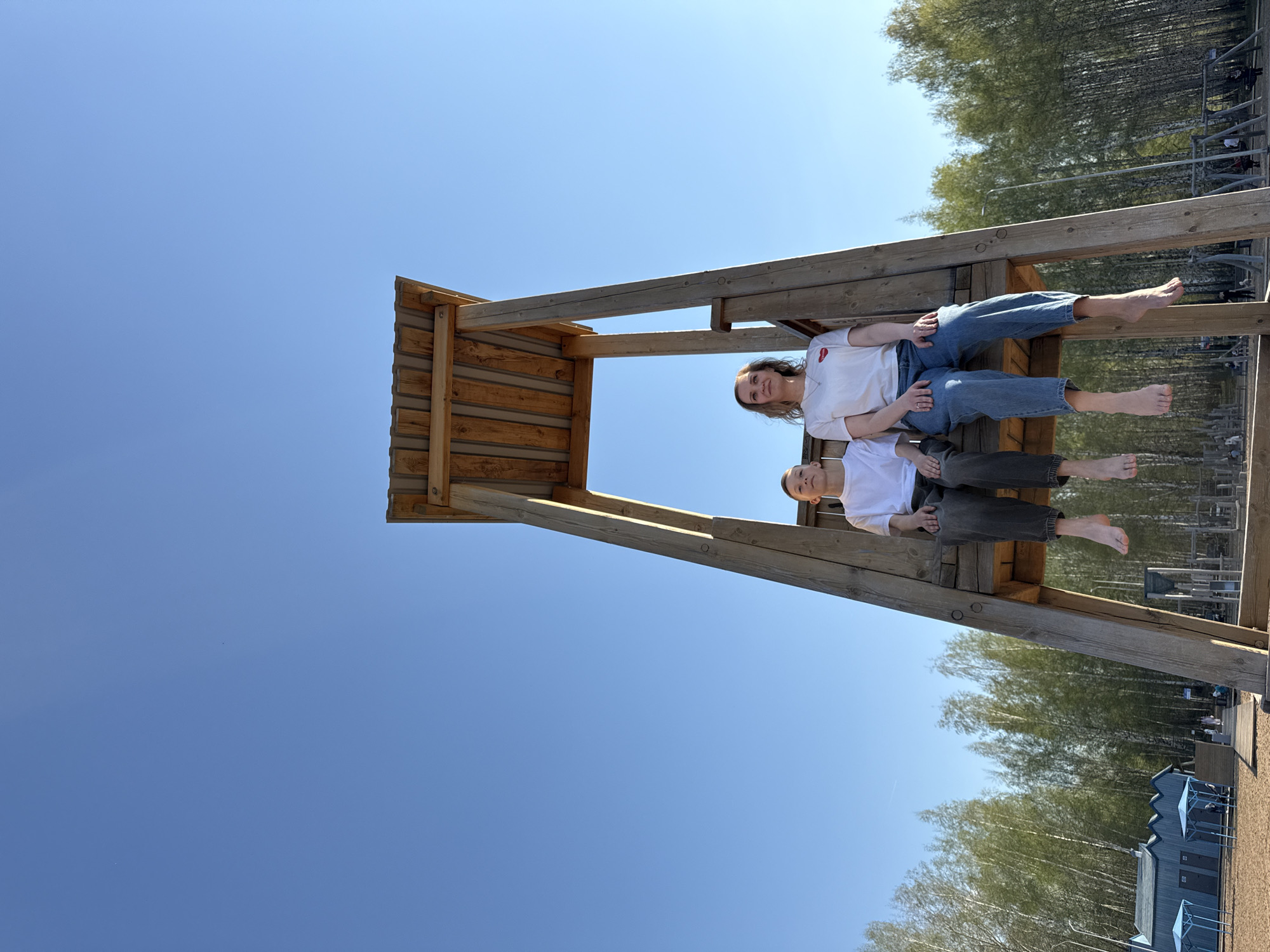 Two individuals, a woman and a child, are situated on a wooden structure. The woman is in a white t-shirt with a red design and jeans. The child is in a white t-shirt and gray pants. Both are barefoot.
The structure appears to be a wooden lookout or platform, potentially part of a playground. The sky is visible, with trees and buildings in the background.
The location is Chaikovsky, Russia.