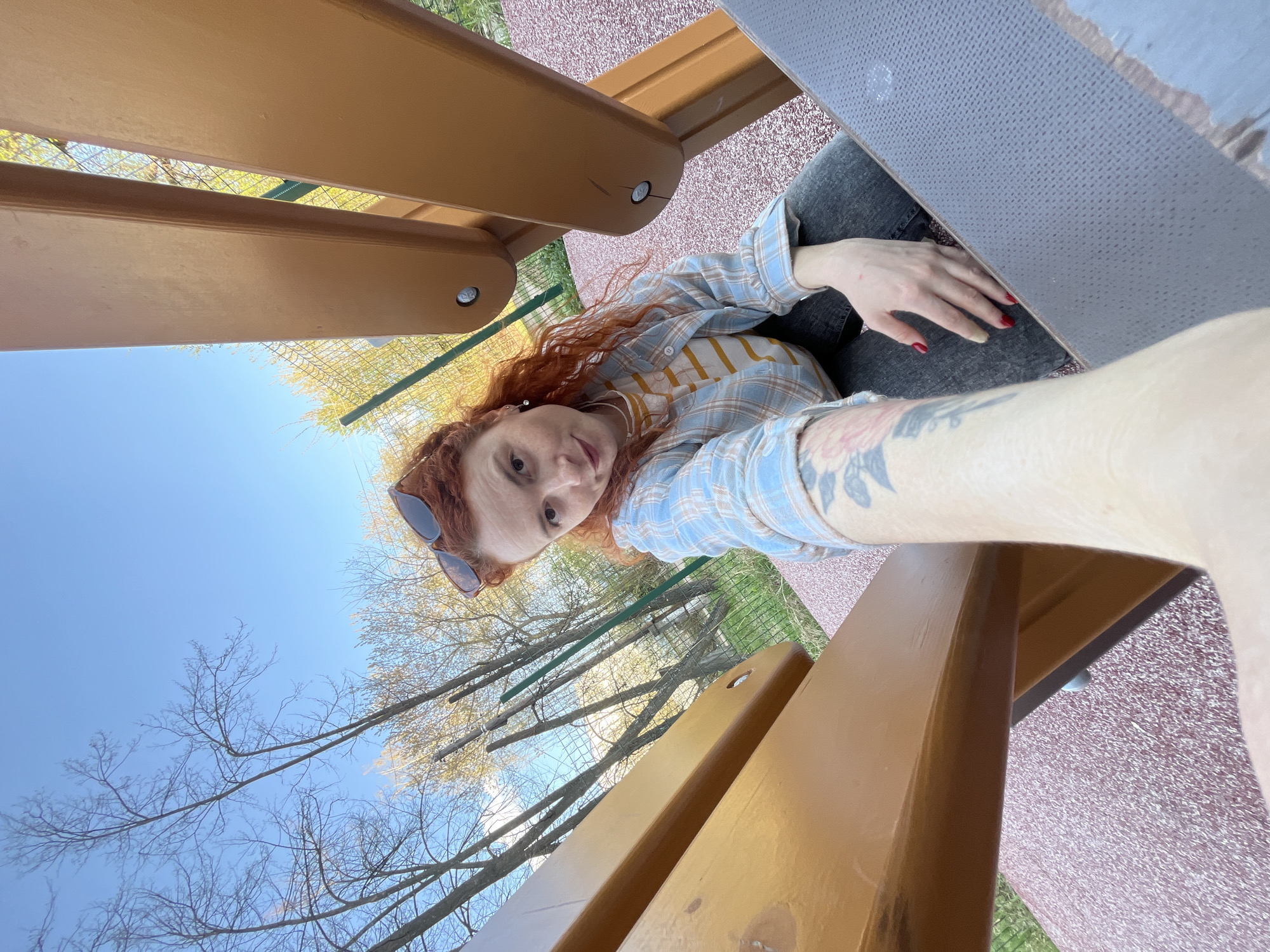 A woman with red hair and sunglasses is sitting at a picnic table in a park, possibly in Chaikovsky, Russia. She is wearing a light blue and white plaid shirt with a yellow and white striped shirt underneath. She has a tattoo on her left arm. The picnic table is made of wood and has a gray concrete top. The ground is covered in a red and white speckled material. Trees and blue sky are visible in the background.